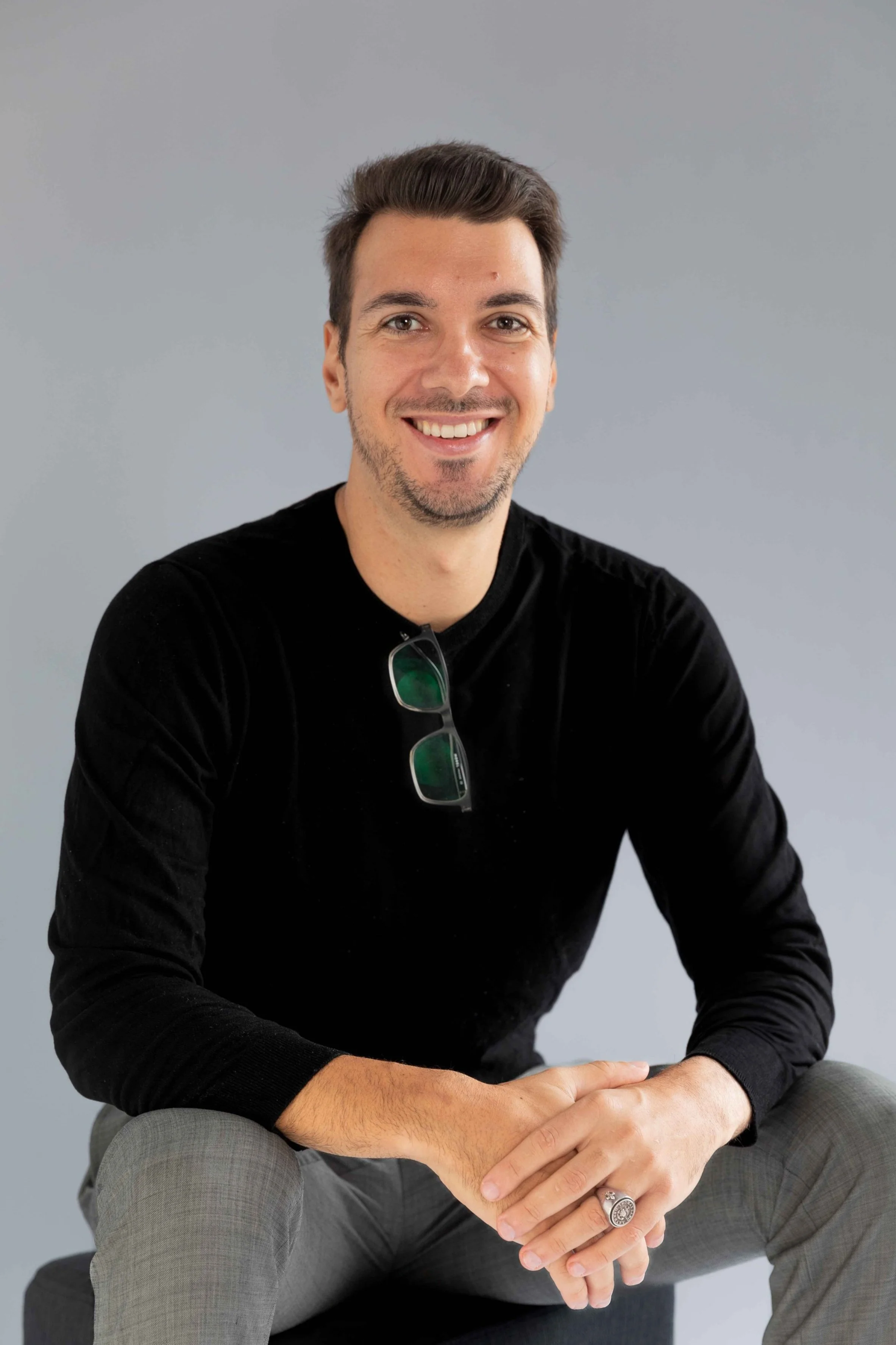 Smiling man wearing a black sweater, sunglasses hanging from the neckline, sitting with hands clasped, against a gray background.