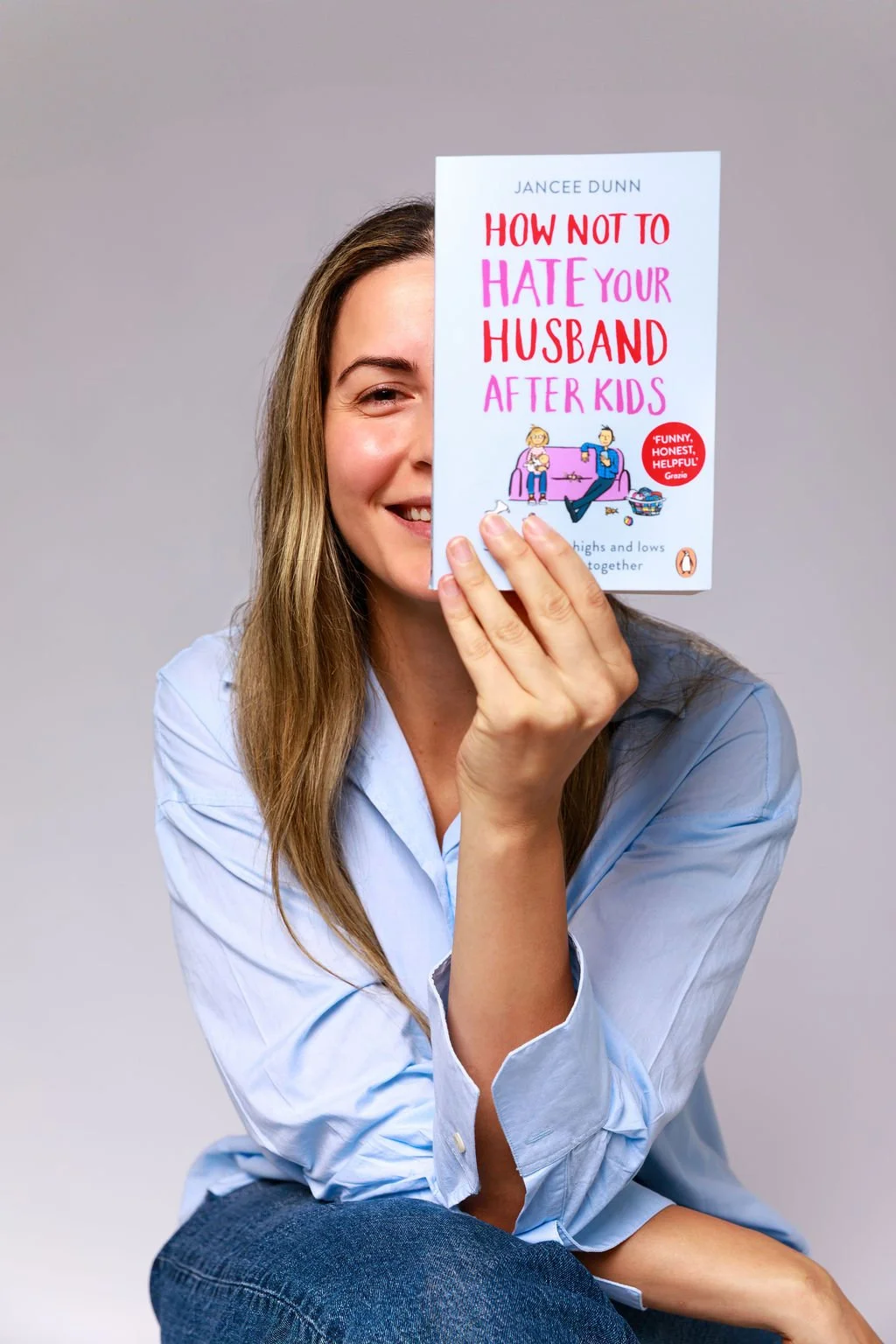 A smiling woman with long hair, wearing a light blue shirt, holding a book titled 'How Not to Hate Your Husband After Kids' in front of her face, partially covering it.