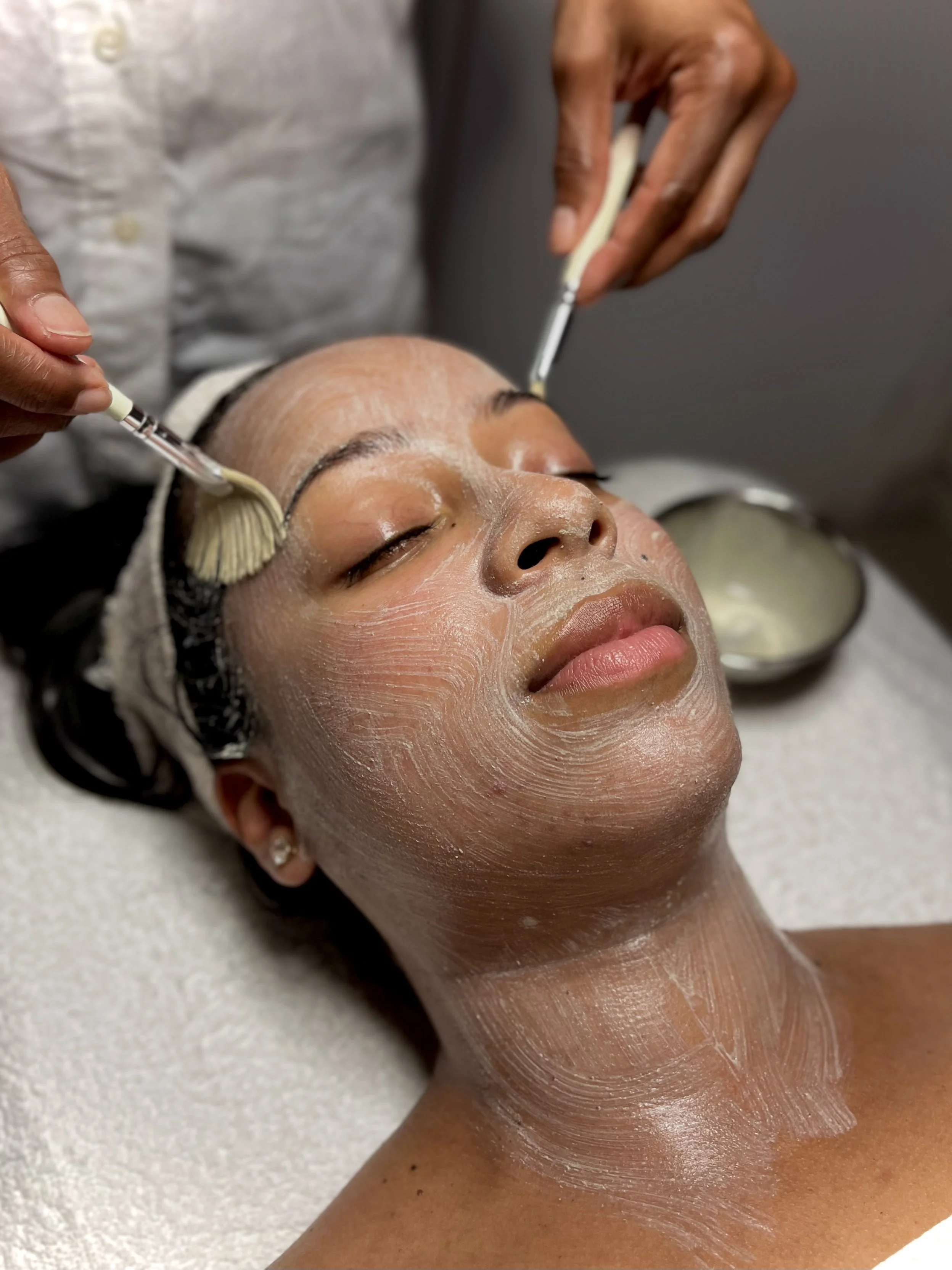 Person receiving a facial treatment, with face and neck covered in a white mask, and two hands applying product with brushes.