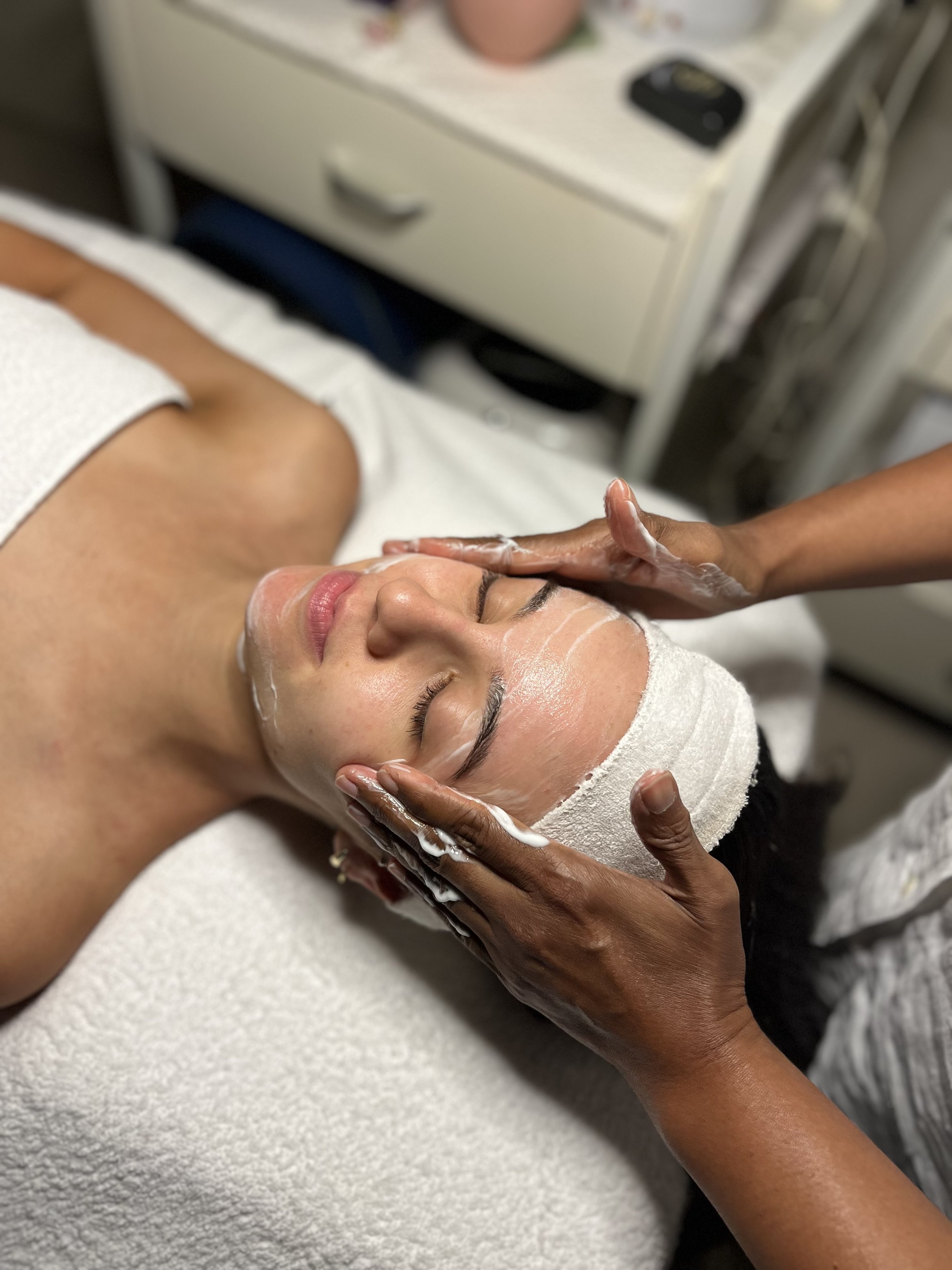 Person receiving a facial treatment, cream applied, therapist's hands massaging face, headband used, spa setting.