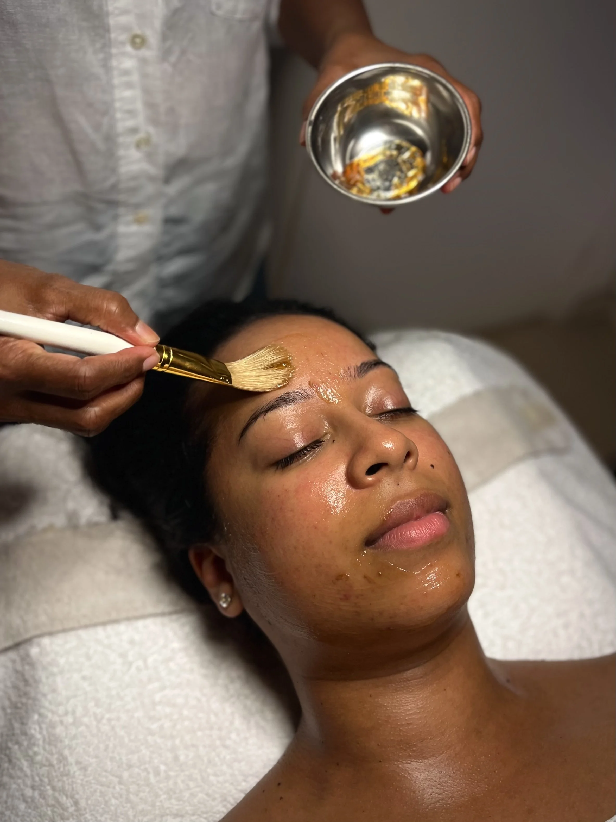 A person receiving a facial treatment with a brush applied to their forehead, while lying on a treatment table with eyes closed. A bowl is held nearby containing a substance used in the facial.