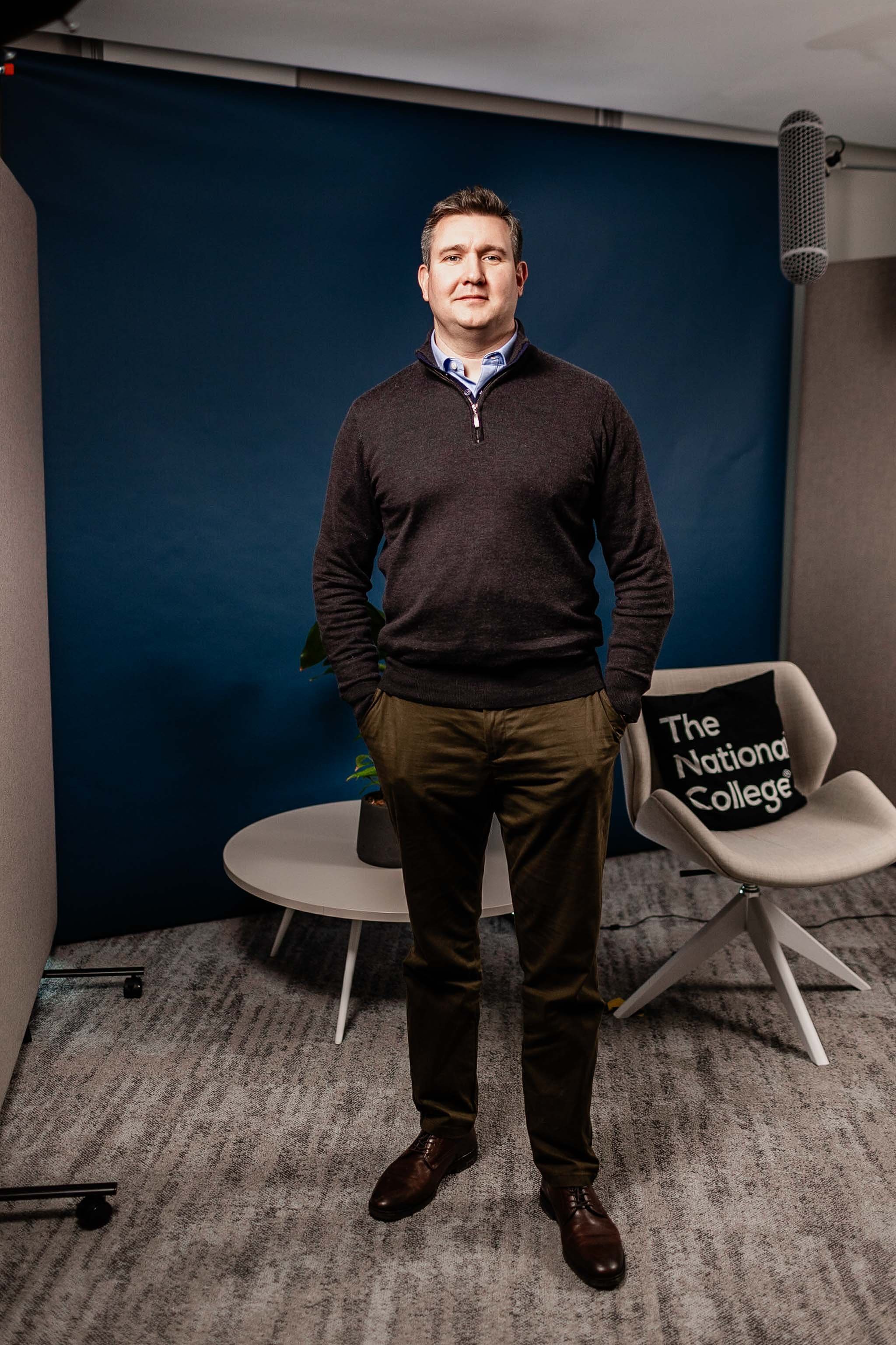 A man standing in a modern office setting with a blue wall backdrop. He is wearing a dark sweater over a collared shirt, with brown pants and dress shoes. There is a round white table and an office chair with a "The National College" pillow on it. Sh