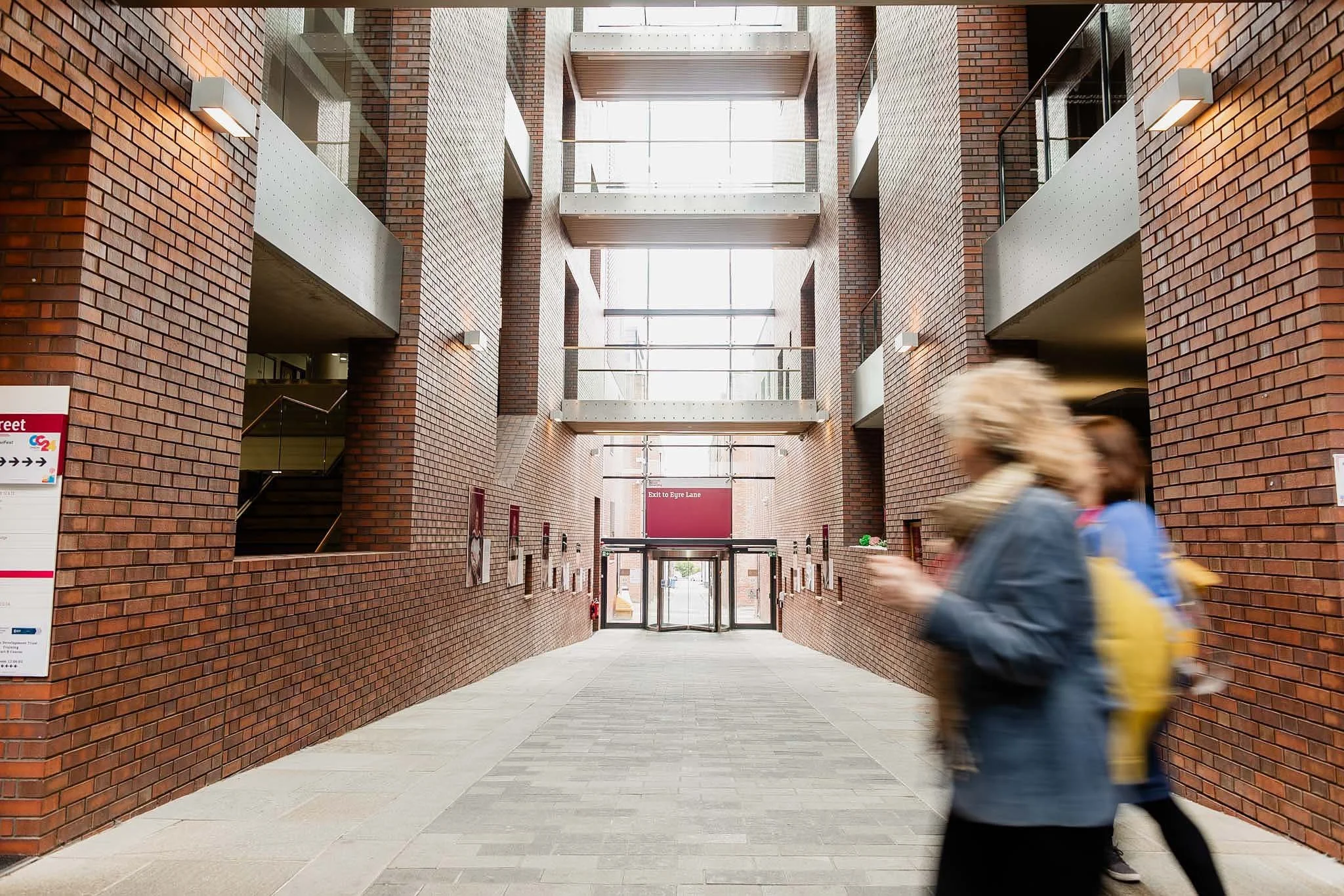 Interior view of a modern building with brick walls, multiple floors, and glass balconies. Two blurred people walk in the foreground, carrying bags. Signs indicate exit directions and nearby streets.