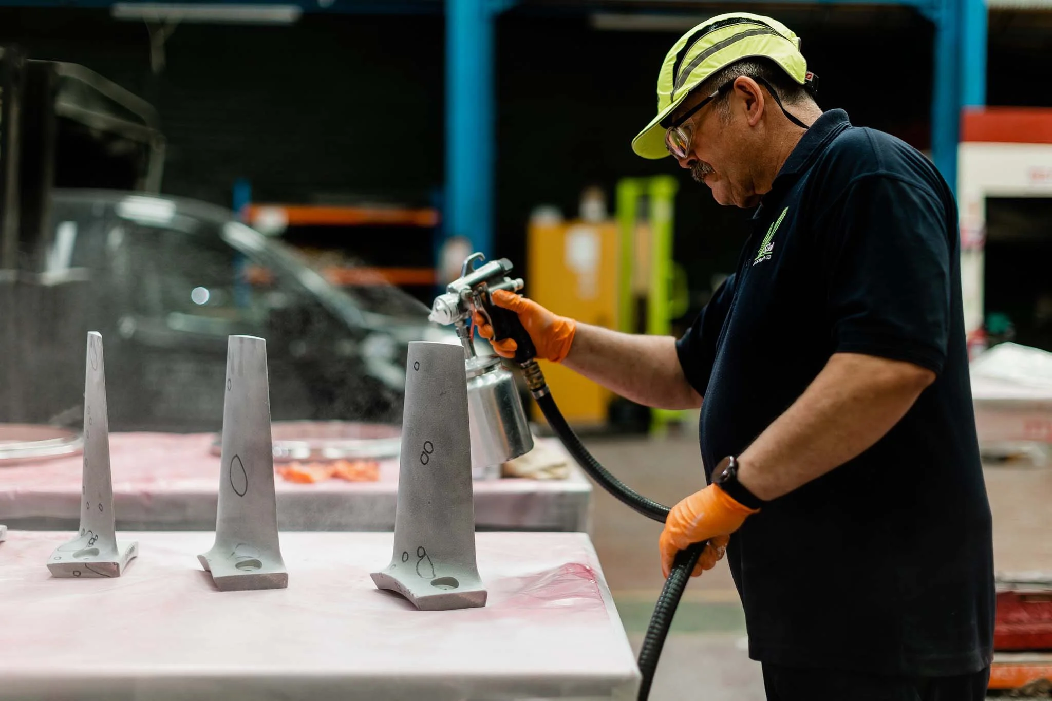 A man wearing protective goggles, gloves, and a reflective helmet sprays a liquid onto paper-mâché molds arranged on a table in a workshop.
