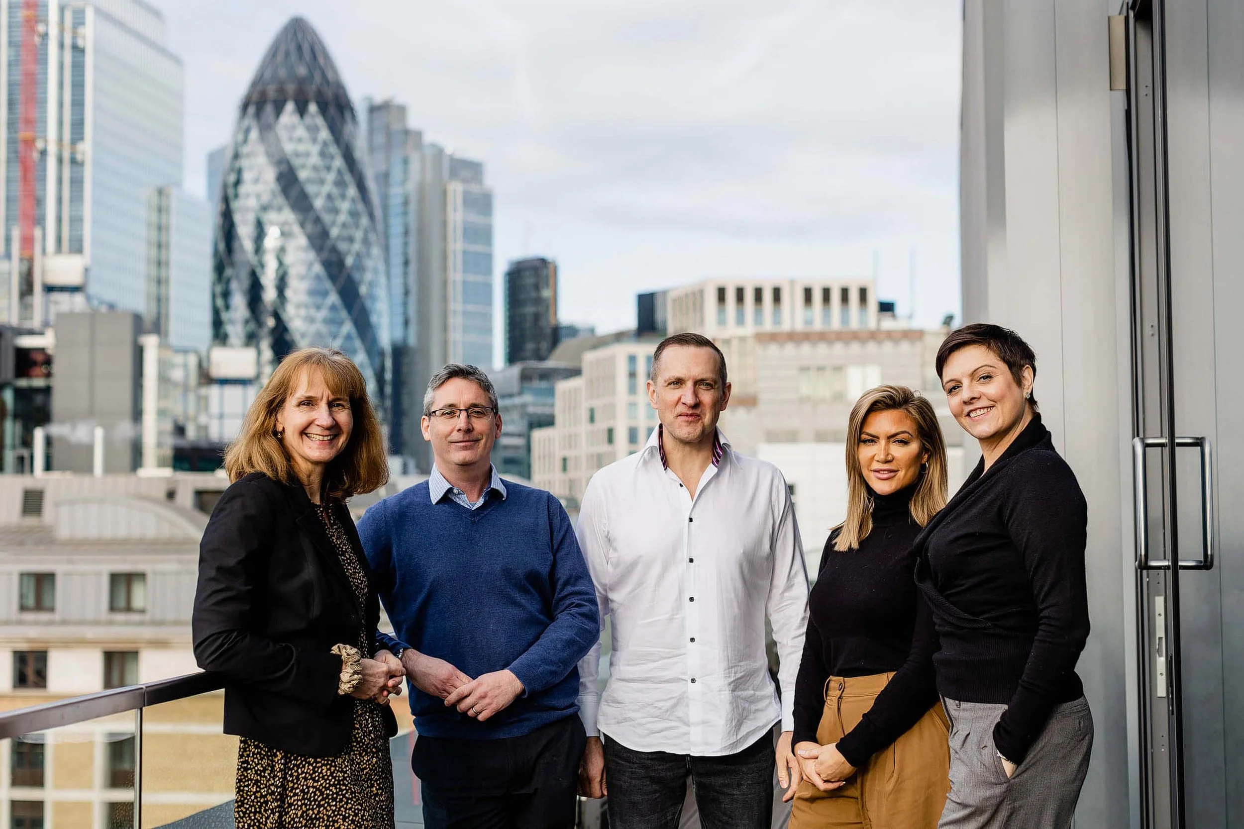 Group of five diverse professionals standing on a balcony of a modern office building with a cityscape skyline including the Gherkin building behind them.
