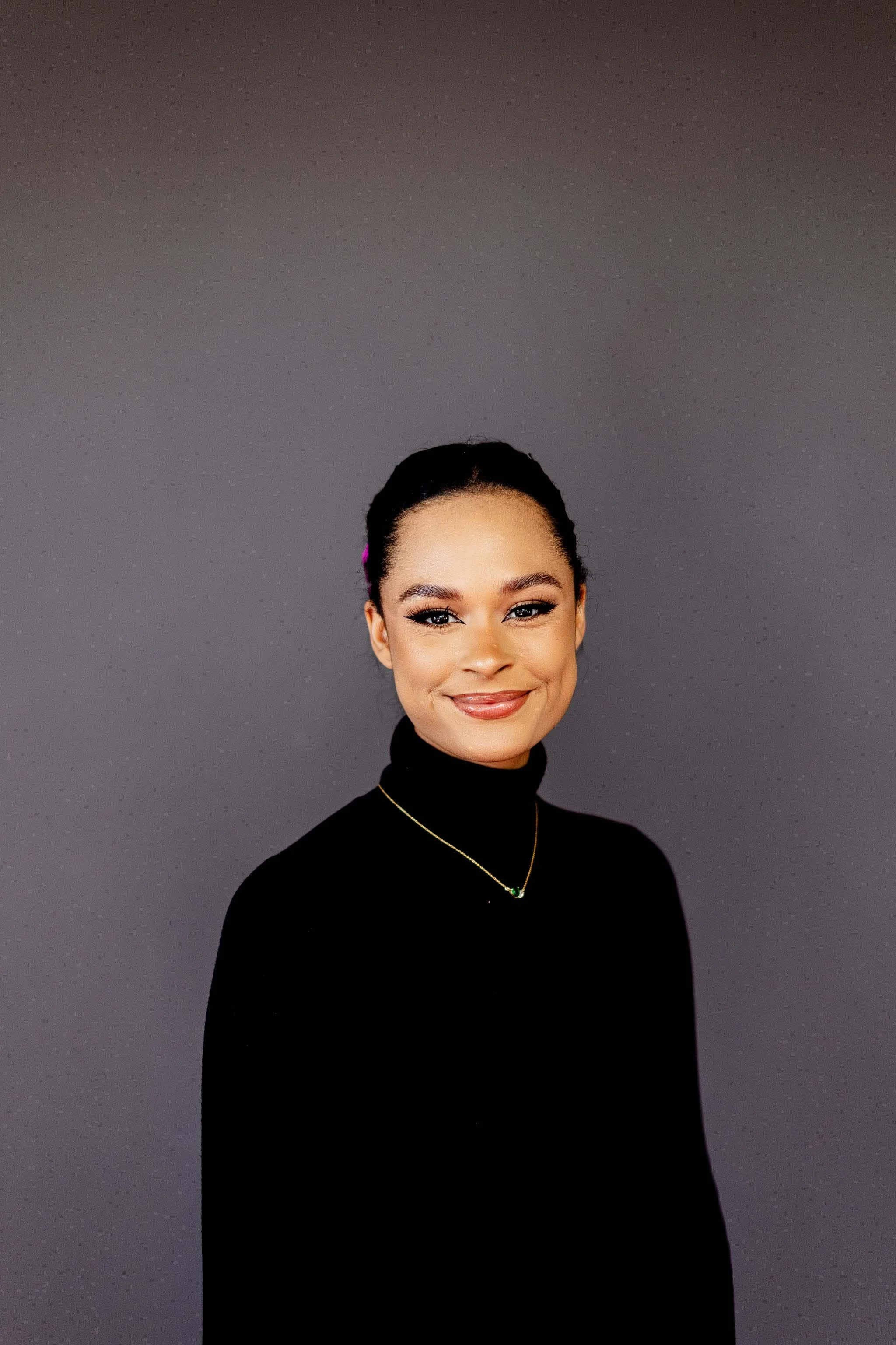 Portrait of a young woman with dark hair pulled back, wearing a black turtleneck, gold necklace with a green pendant, and pink hair clip, smiling against a gray background.