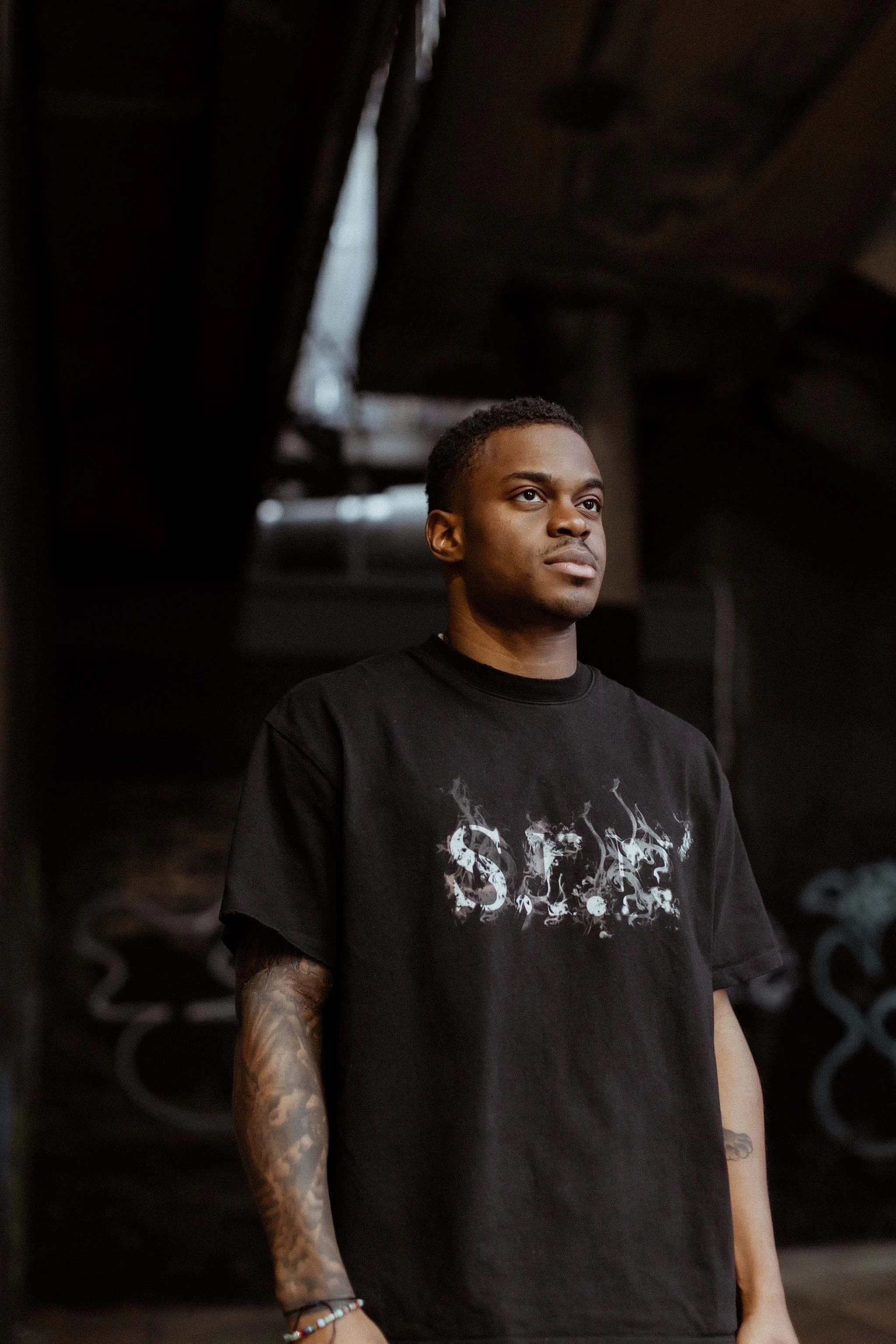 A young man with dark skin, short curly hair, and tattoos on his right arm stands indoors, wearing a black T-shirt with a graphic design, looking off to the side with a serious expression.