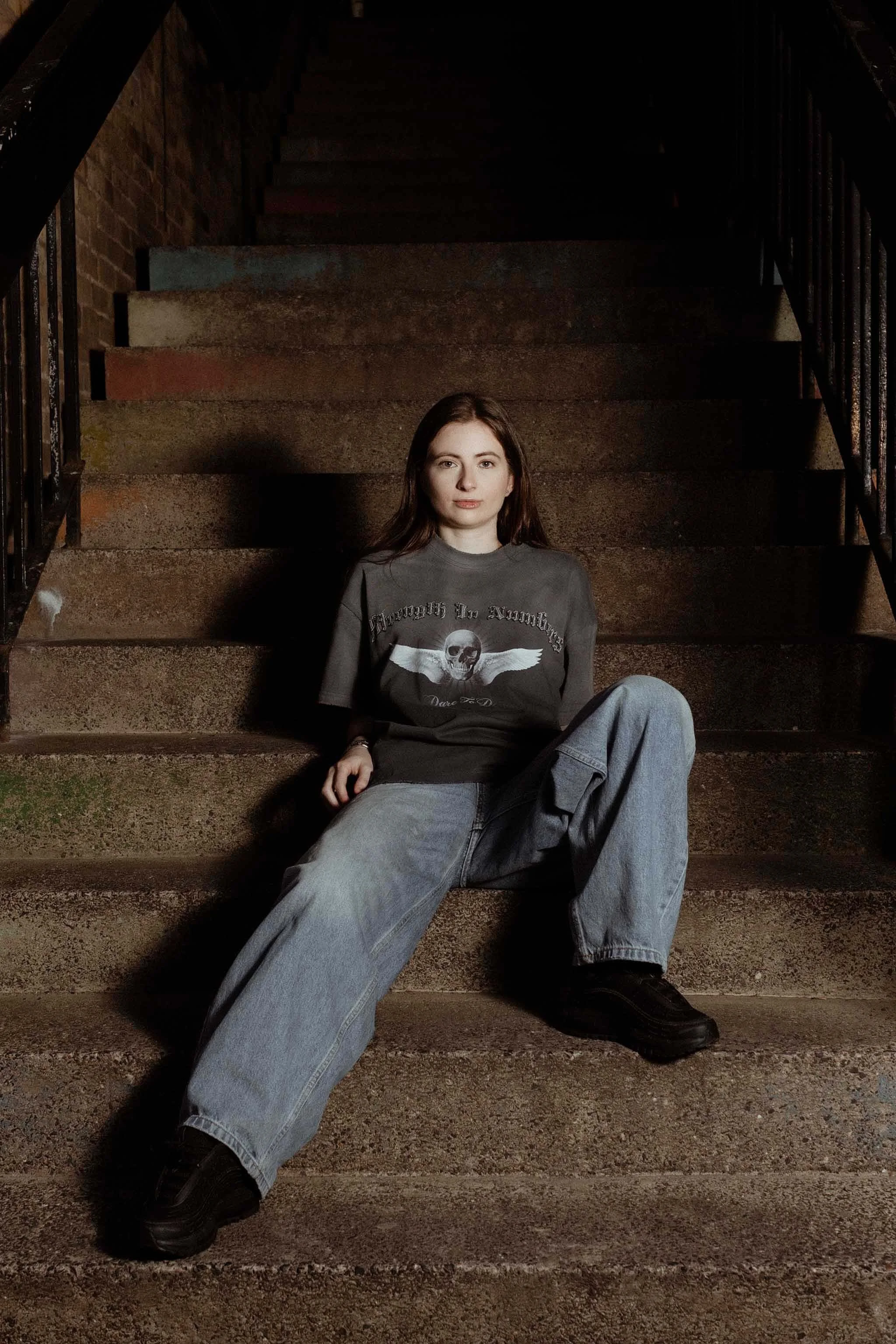 A young woman with long brown hair sitting on outdoor concrete stairs at night, wearing a dark graphic T-shirt, baggy jeans, and black shoes.