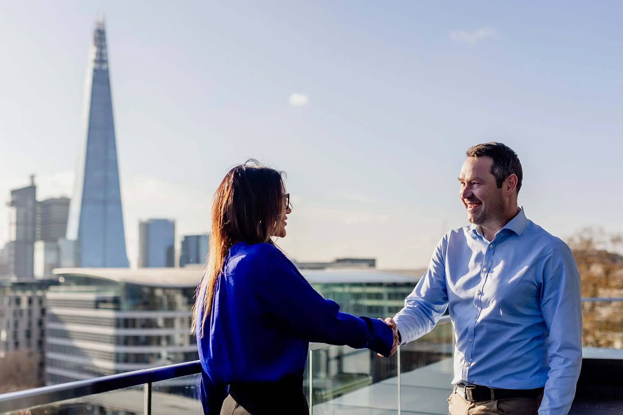 A man and a woman shaking hands on a rooftop in a city with tall buildings, including the Salesforce Tower, during daylight.