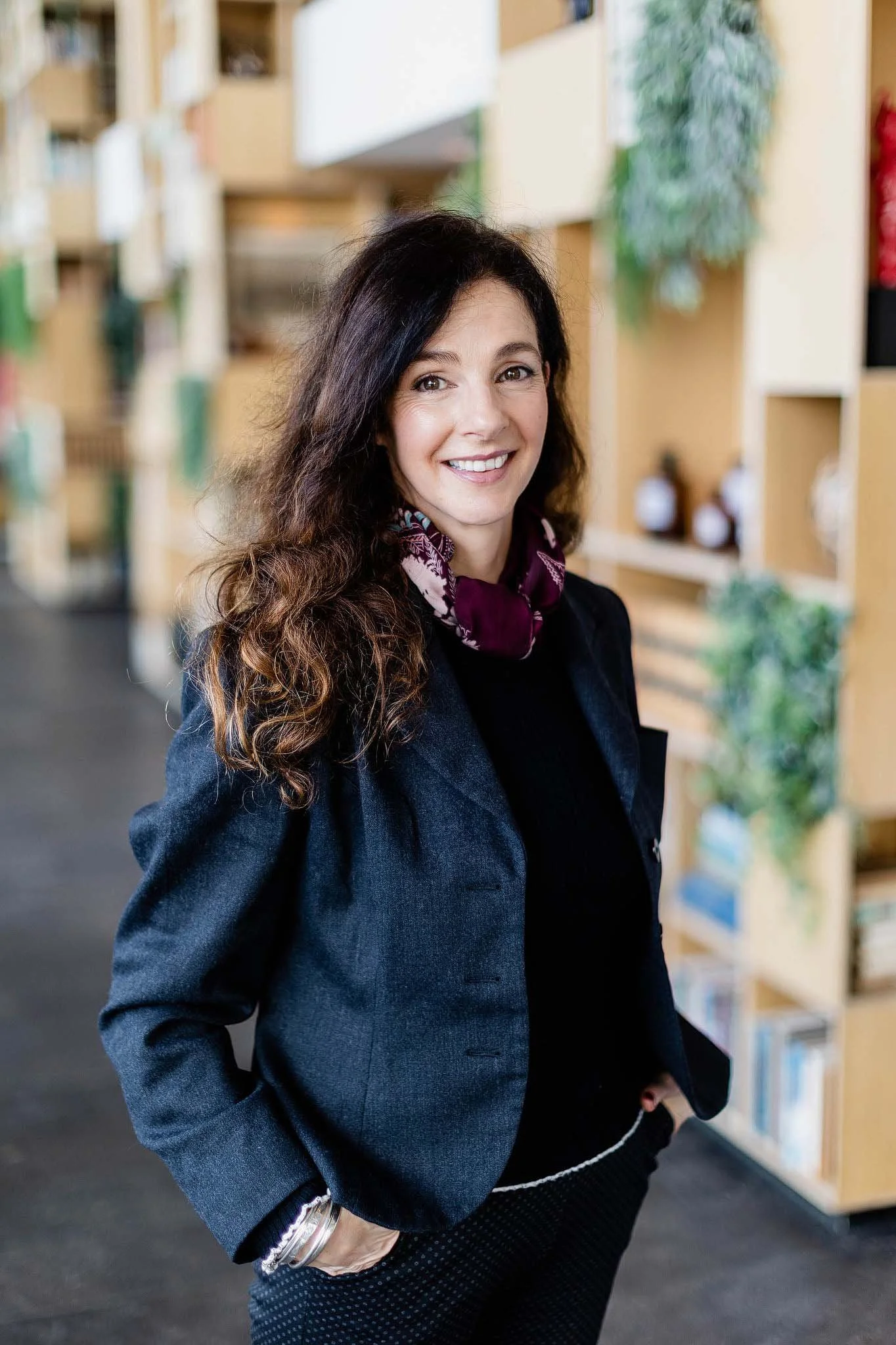 A woman with long, wavy dark brown hair, smiling, wearing a dark blazer, polka-dot pants, and a patterned scarf, standing in front of a bookcase in a library or bookstore.