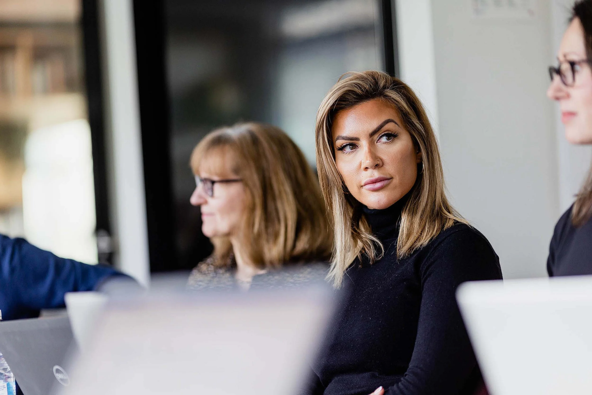 Three women sitting at a table in a professional setting, engaged in a discussion. The woman in focus has long brown hair, light skin, and is wearing a black top. The background shows a blurred office environment.