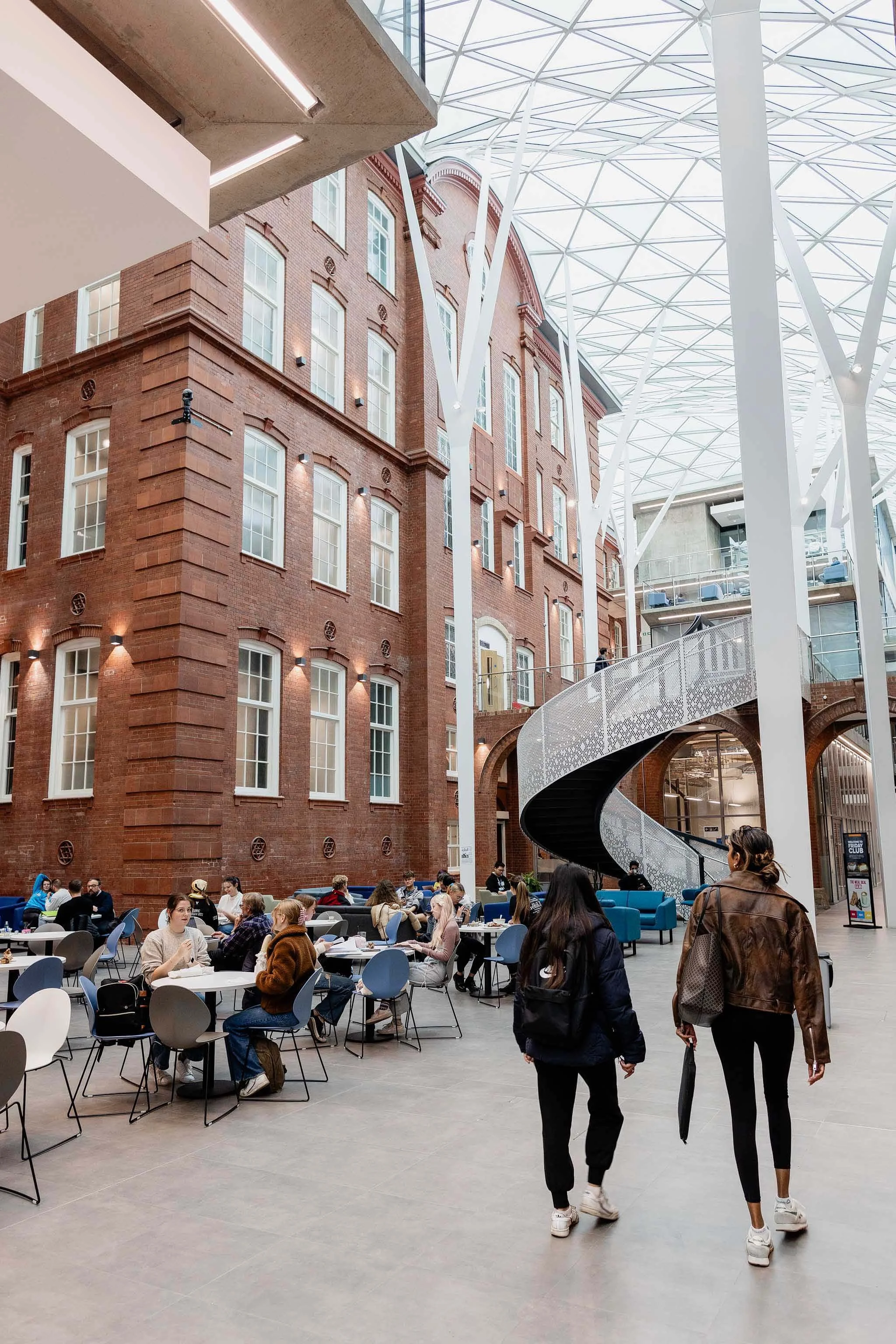 Interior of a modern building with a glass ceiling, brick walls, and a dining area filled with people sitting at tables. Two women walk past in the foreground.