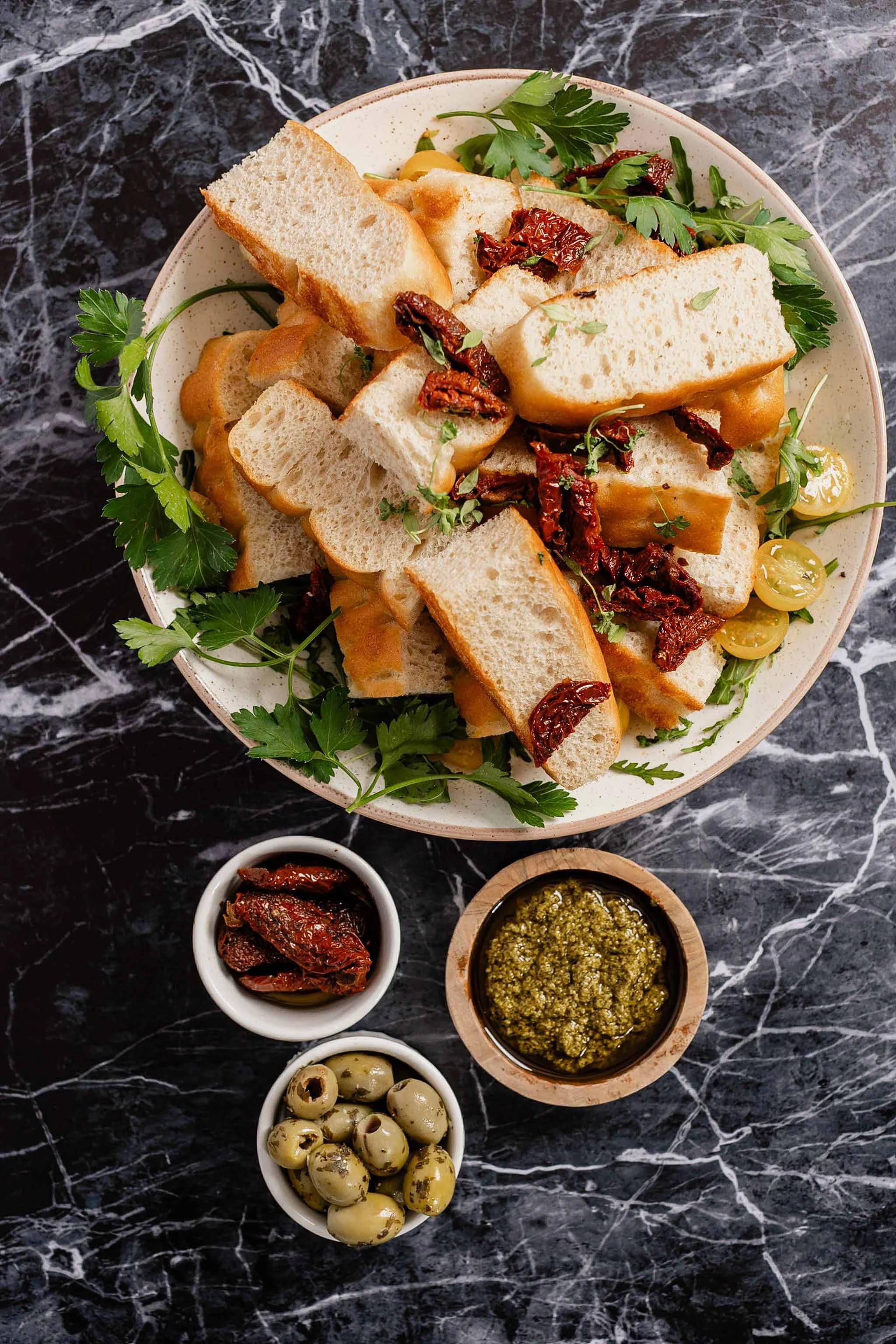 A round beige bowl containing sliced baguette toasts, sun-dried tomatoes, and fresh parsley leaves on a dark marble surface. Surrounding the bowl are small bowls with green olives, sun-dried tomatoes, and green pesto sauce.