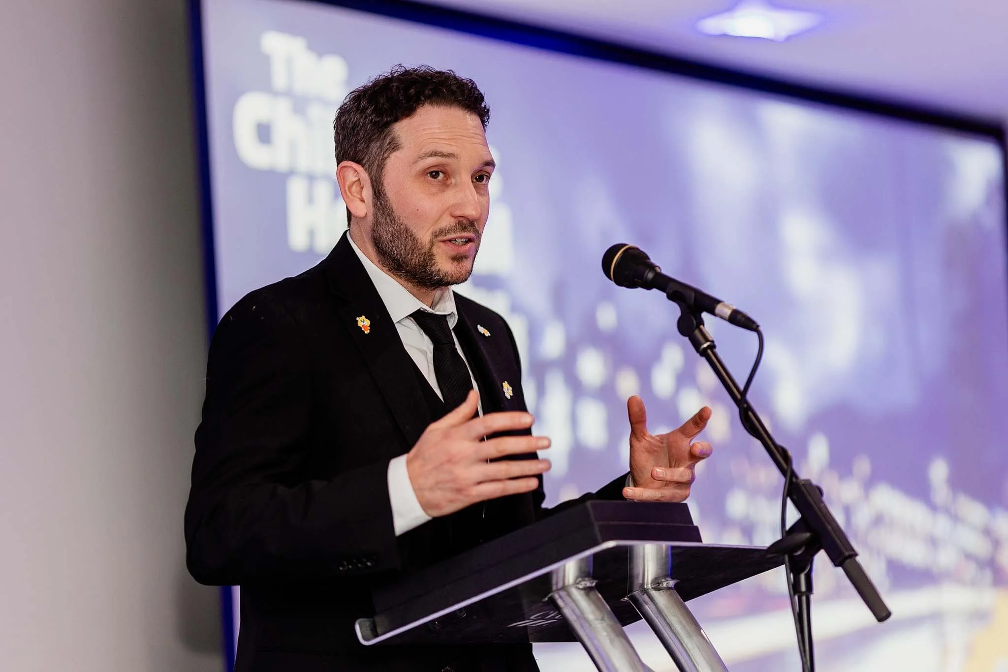 A man with dark curly hair and a beard, dressed in a black suit and white shirt, speaking into a microphone at a podium during a presentation or conference.