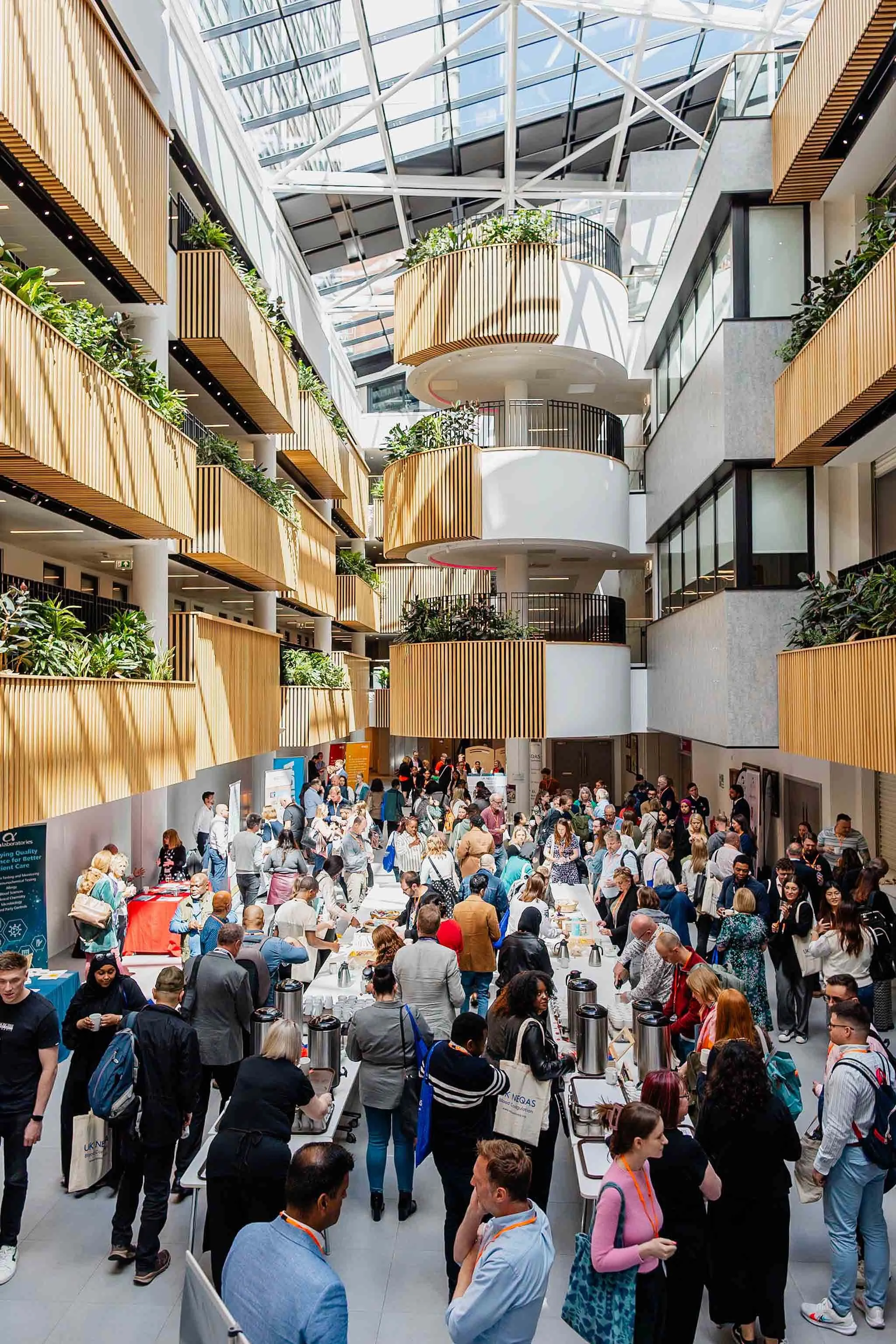 Indoor event with many people gathering around tables with refreshments, in a modern building with glass ceiling and wooden balconies.