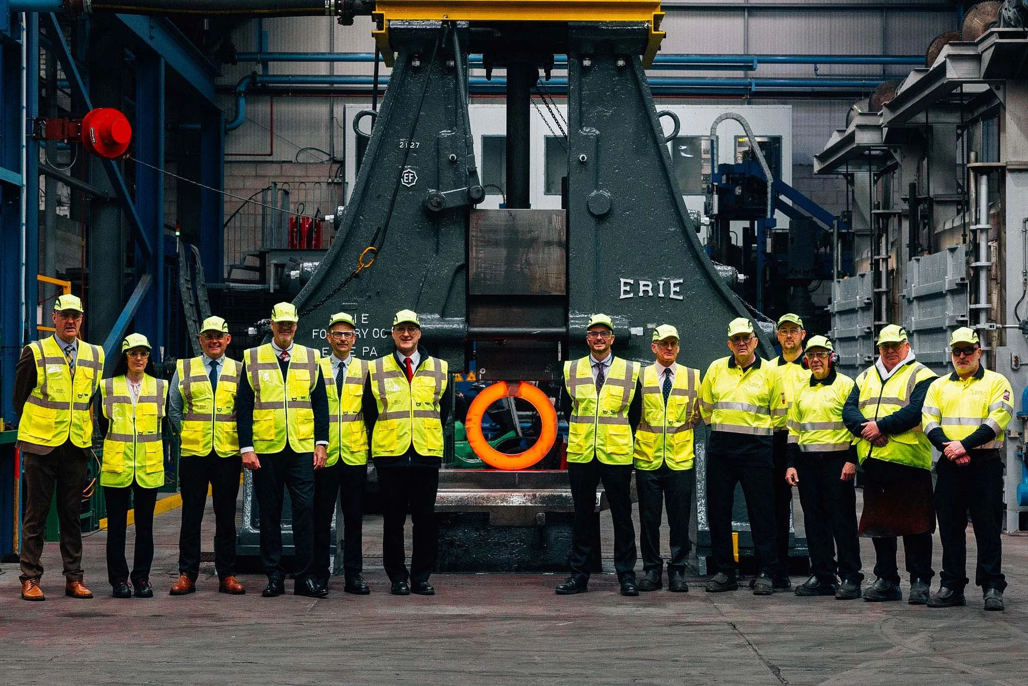 Group of people in safety vests and hard hats standing in front of industrial machinery inside a factory or manufacturing plant.