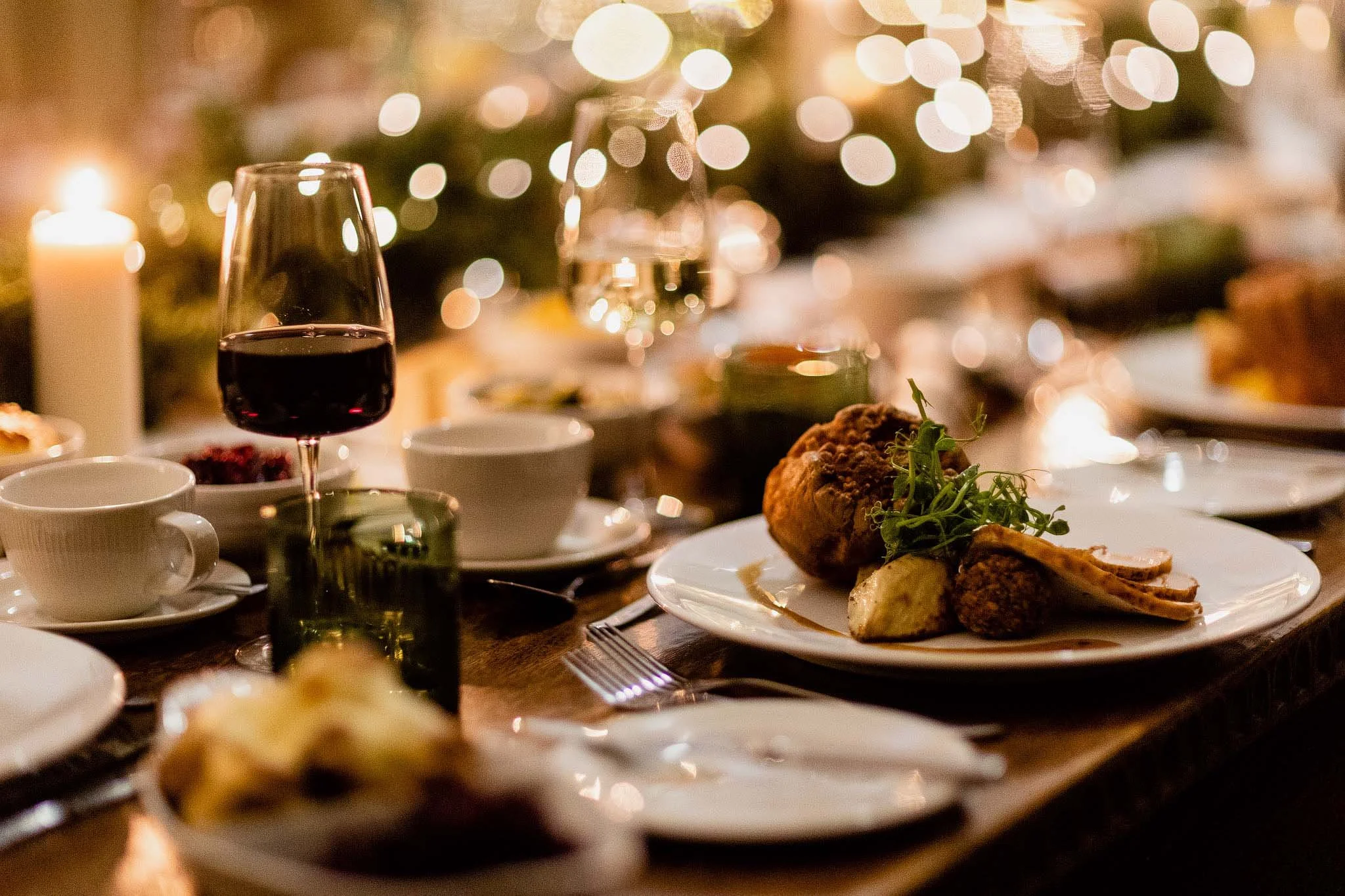 A glass of red wine and a plate with a roast chicken dish on a dining table, with candles and blurred festive string lights in the background.