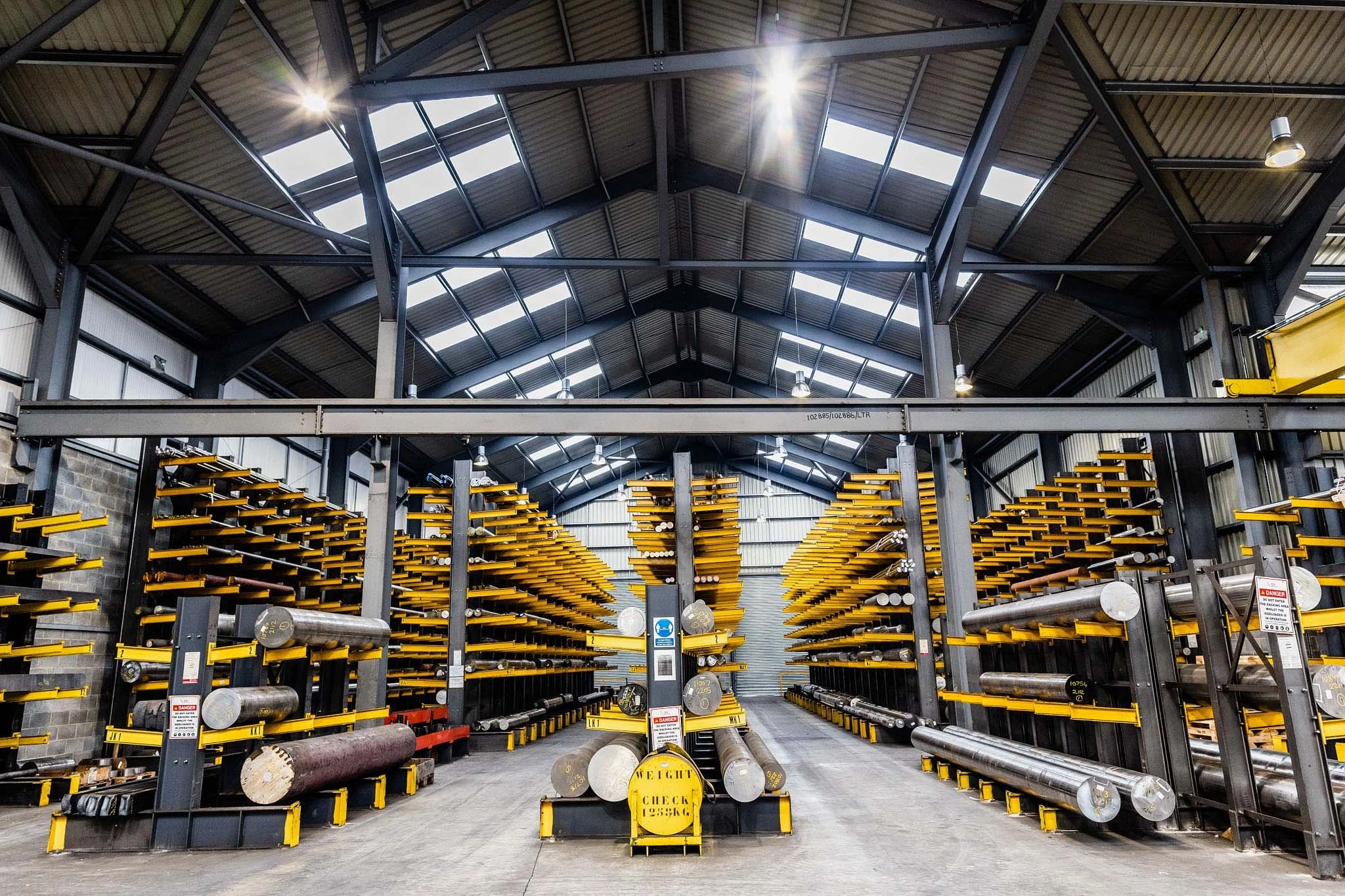 Inside a warehouse with metal racks holding large metal pipes and beams, with a high ceiling and skylights letting in natural light.