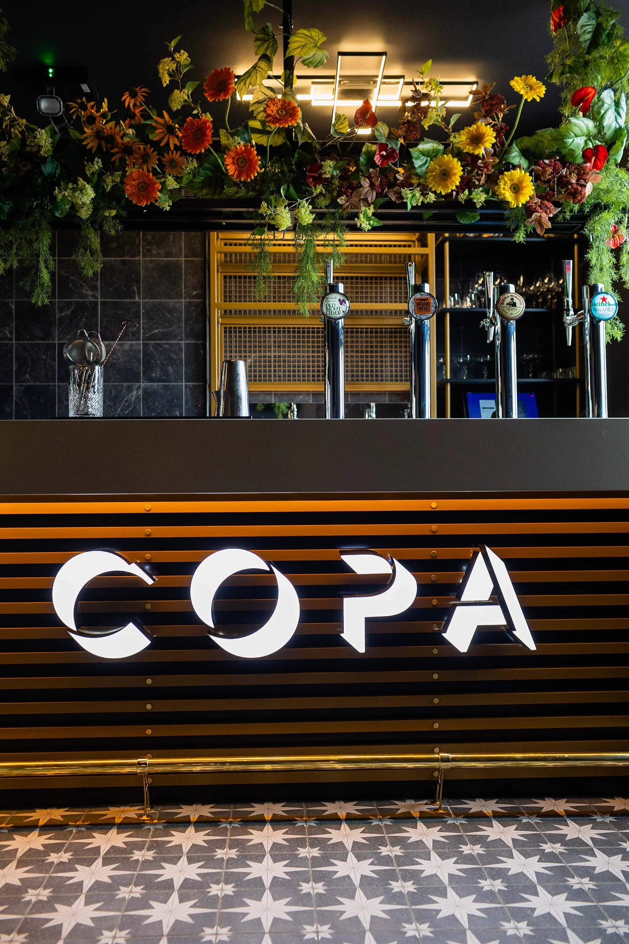 Decorated bar area with flowers and illuminated sign reading 'COPA' on the front, black wall with geometric lighting, patterned floor tiles.