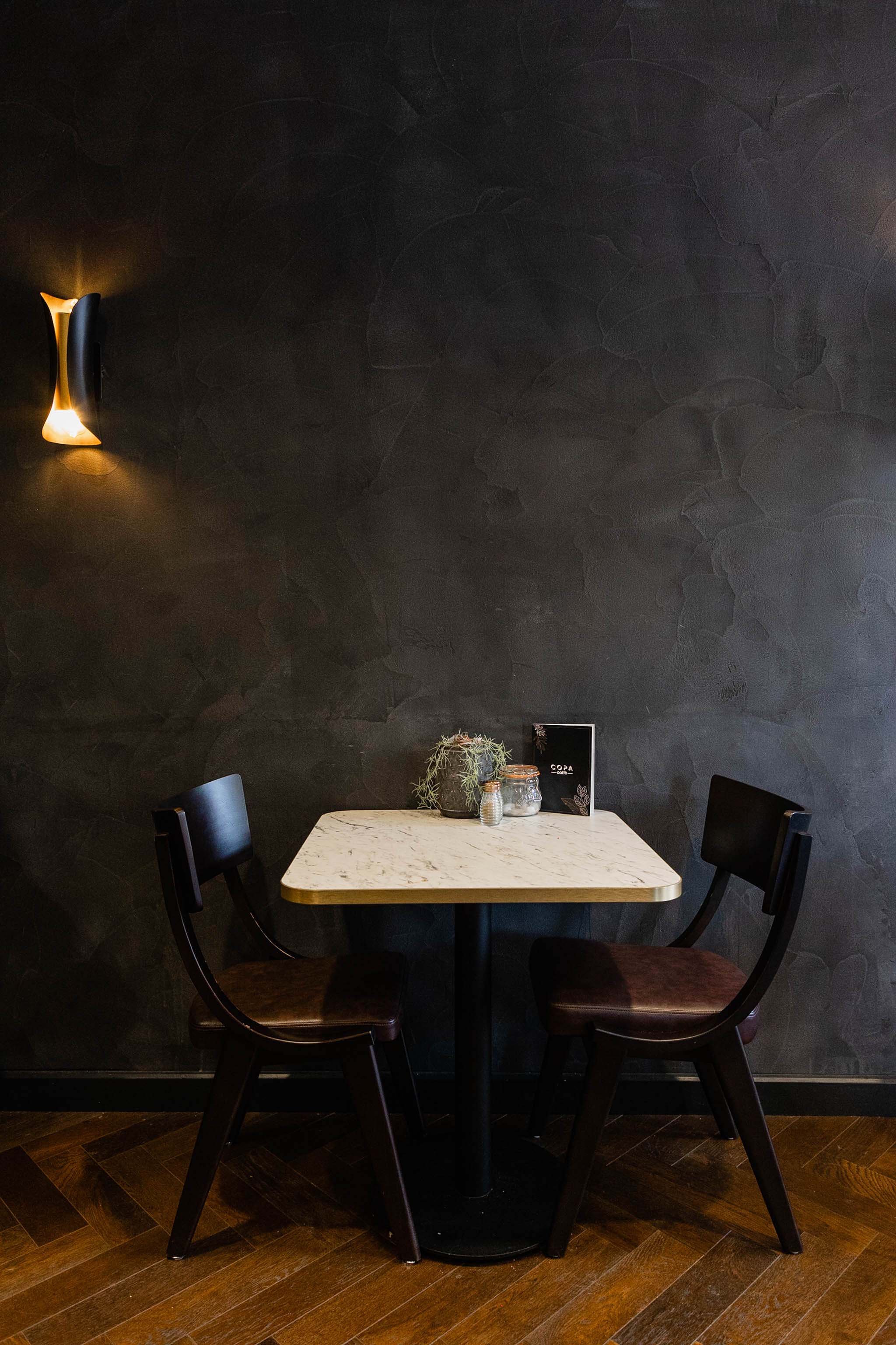 A small round table with a marble top, two black chairs, a plant in a decorative pot, and jars, set against a dark textured wall with a wall-mounted light on the left.