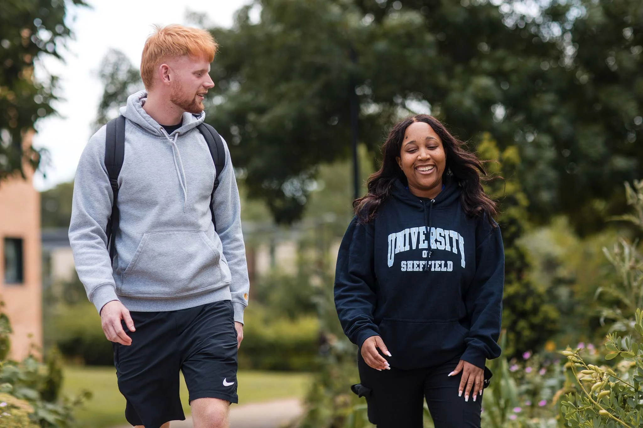 A young man with red hair and a woman with dark hair walk together outdoors in a park, smiling and talking. The woman is wearing a black hoodie with 'University of Sheffield' printed on it. The man has a backpack and grey hoodie. They are surrounded 