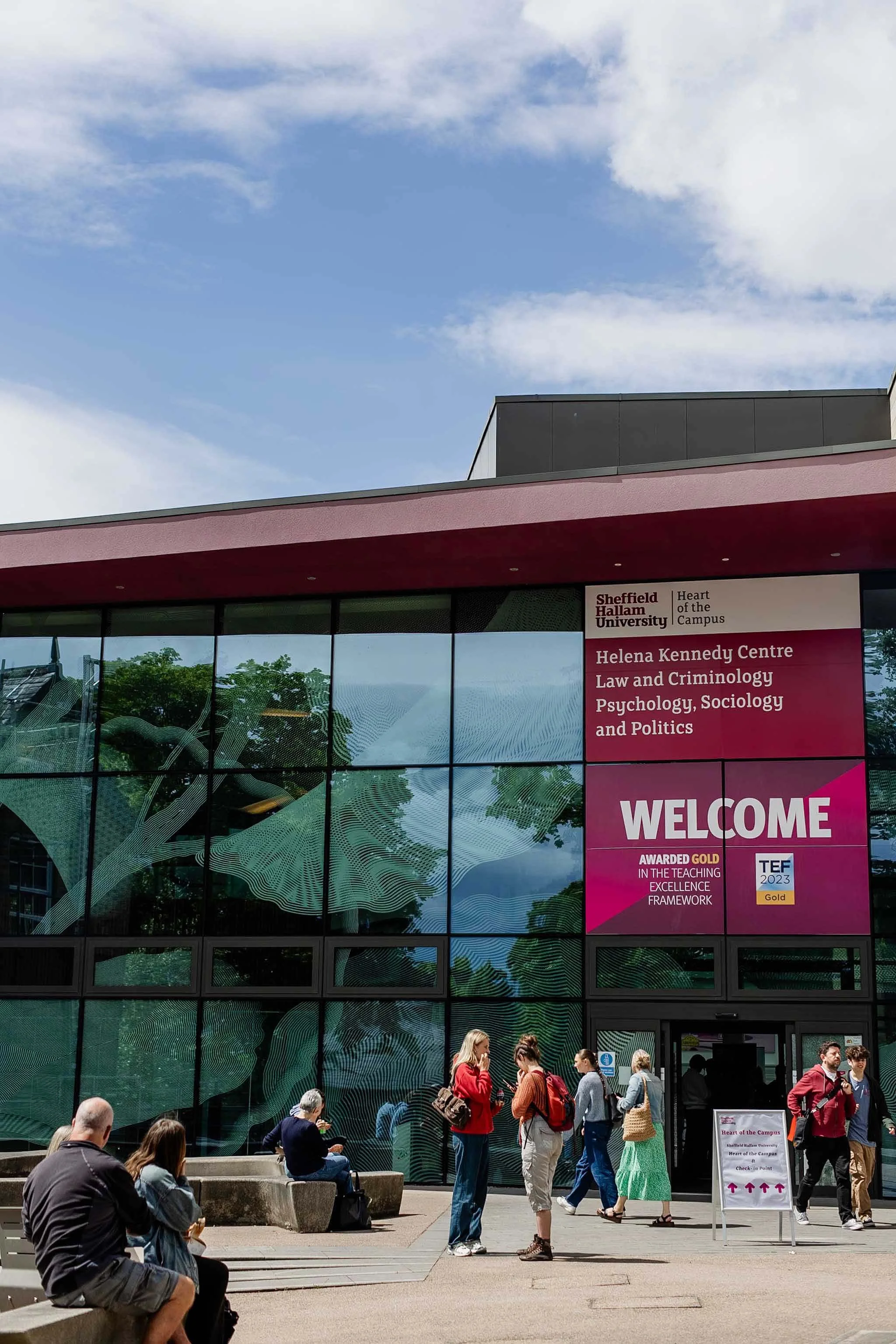 People gathered outside Sheffield Hallam University Helena Kennedy Centre for Law and Criminology, with a modern glass building and a sign welcoming visitors. The scene includes individuals standing, sitting, and walking, some engaging in conversatio
