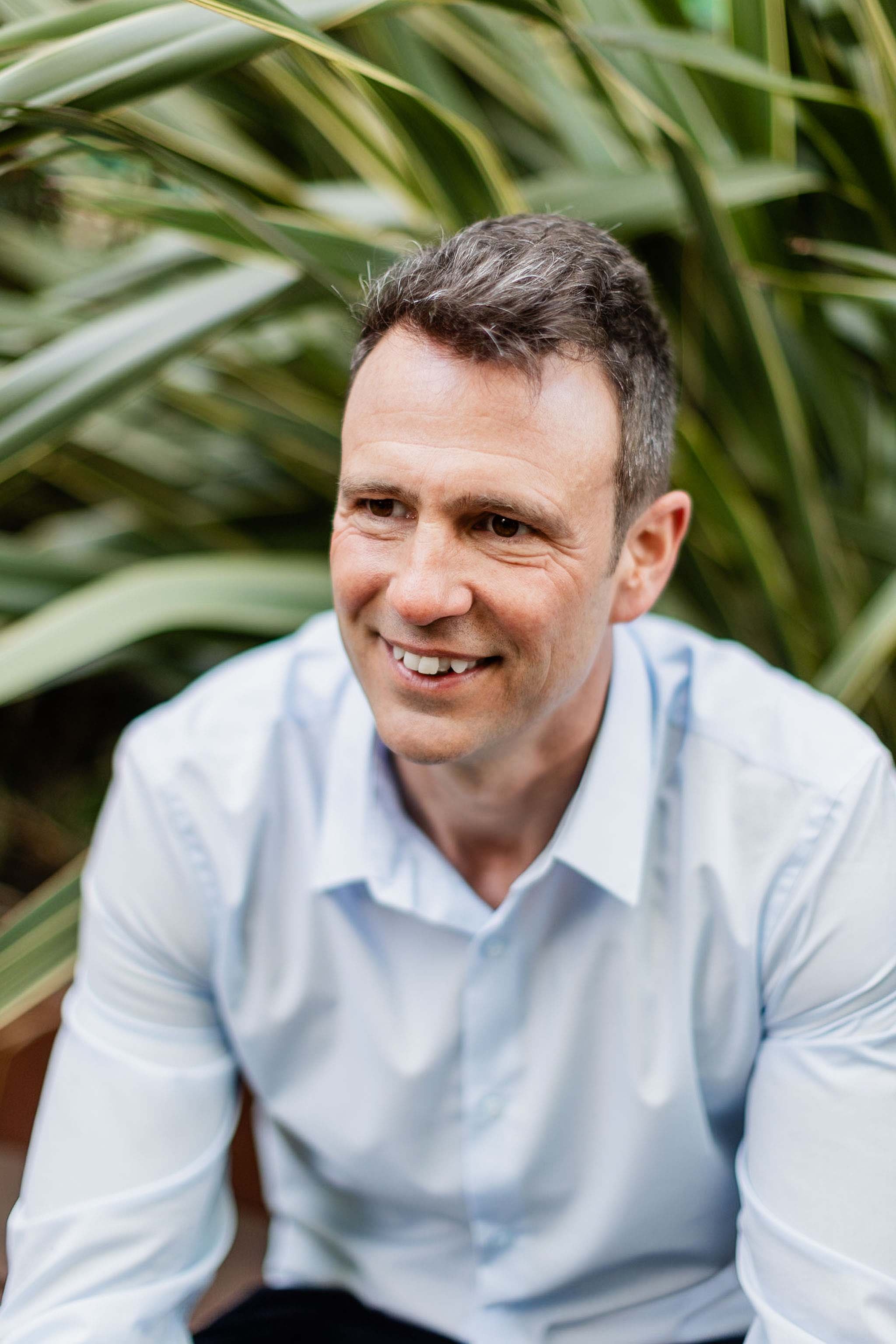 A man with short dark curly hair smiling outdoors surrounded by large green plants.