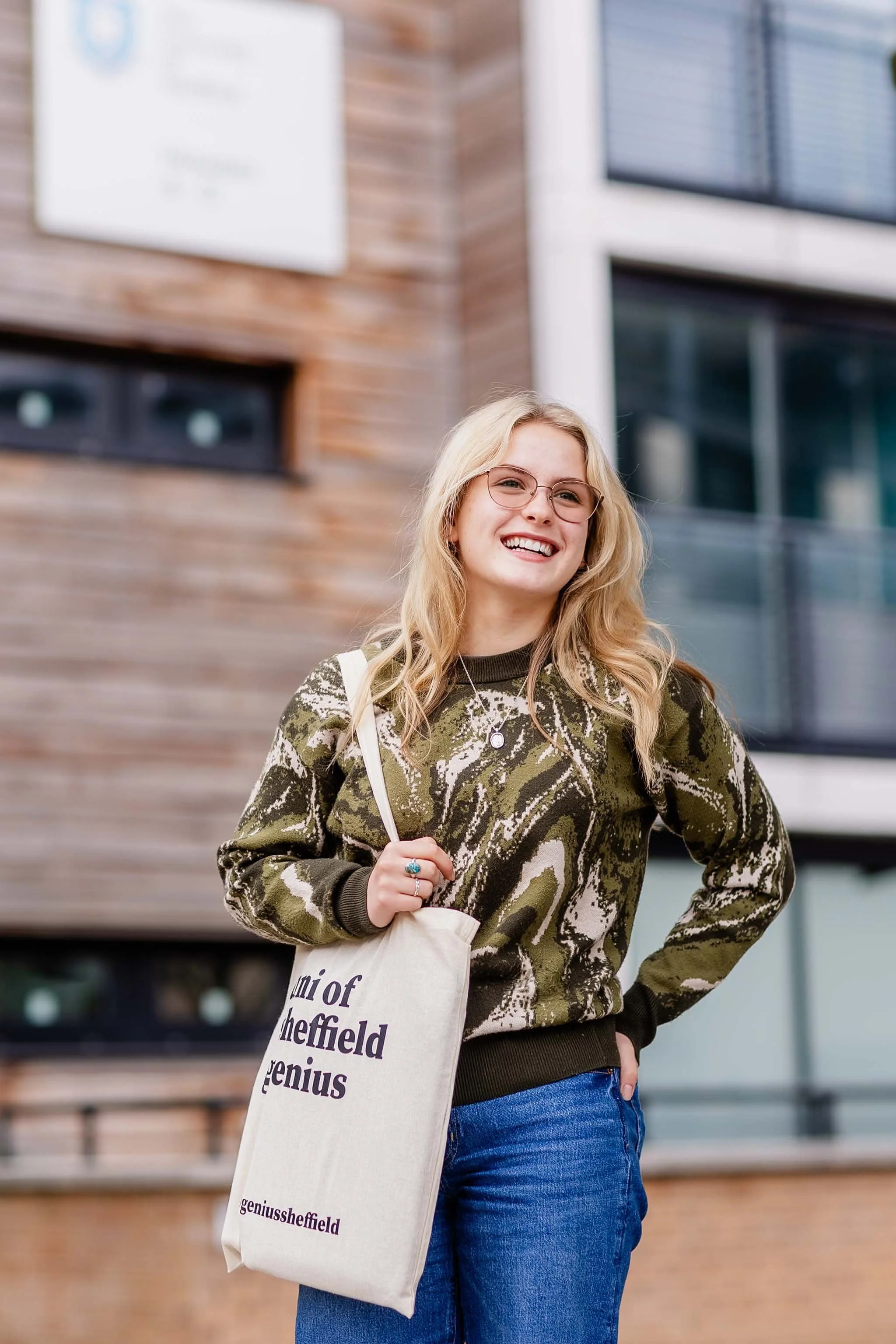 A young woman with long blonde hair, glasses, and a camouflage sweater standing outdoors in front of modern buildings, smiling and holding a beige tote bag that reads 'Genius of Sheffield'.