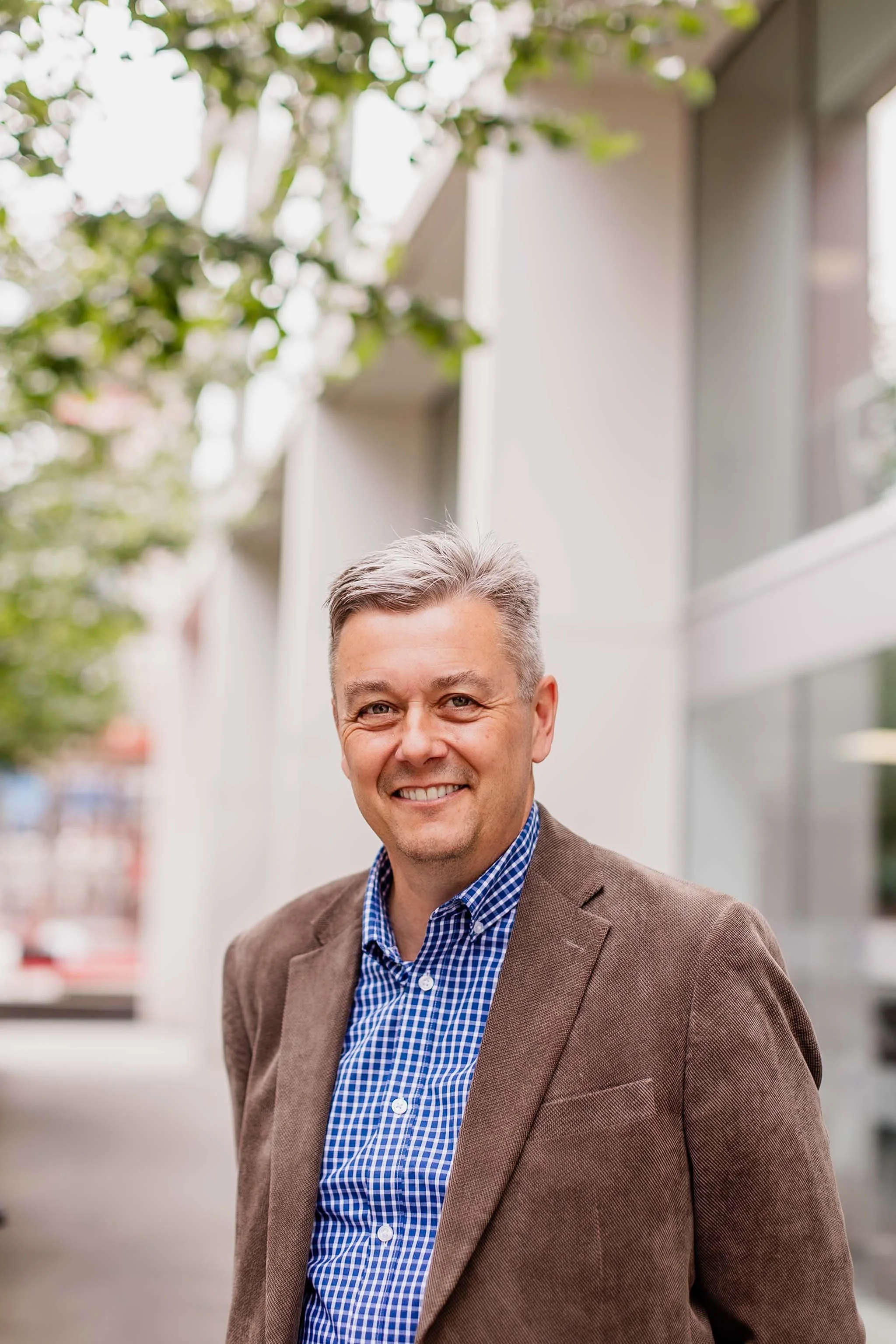 A middle-aged man with gray hair, smiling, wearing a brown blazer and a blue checkered shirt, standing outdoors with trees and a building in the background.