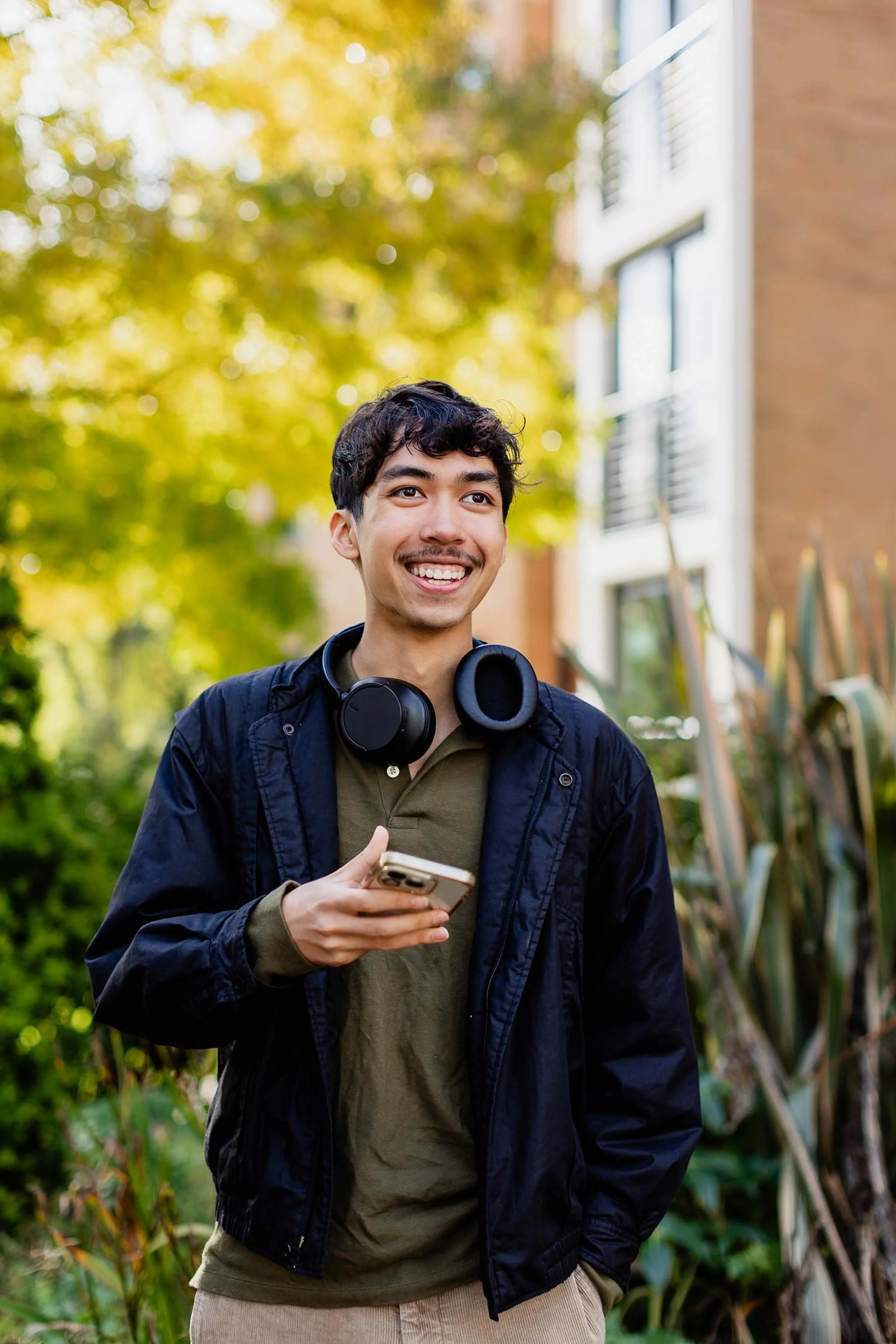 A young man smiling and looking off into the distance while holding a smartphone in his hand. He is outdoors with autumn trees in the background. He is wearing a dark jacket with headphones around his neck and a green shirt underneath.
