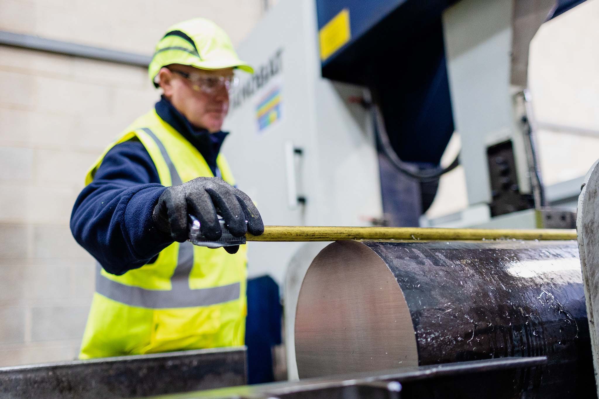 A worker in safety gear, including a yellow reflective vest, gloves, hat, and safety glasses, is operating a large machine in an industrial setting, possibly cutting or shaping a thick cylindrical piece of material.