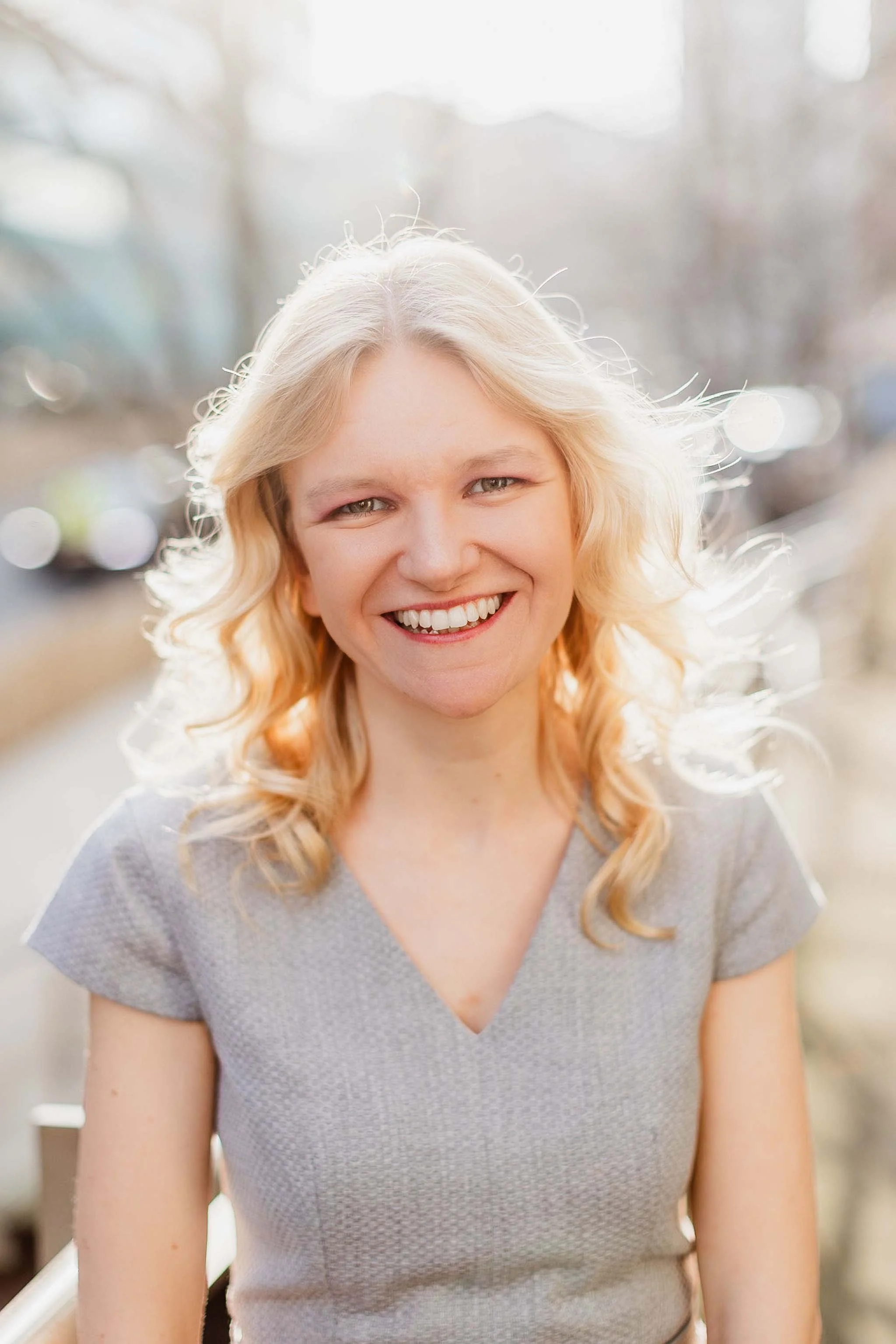 A woman with blonde, wavy hair smiling outdoors on a bright, sunny day.