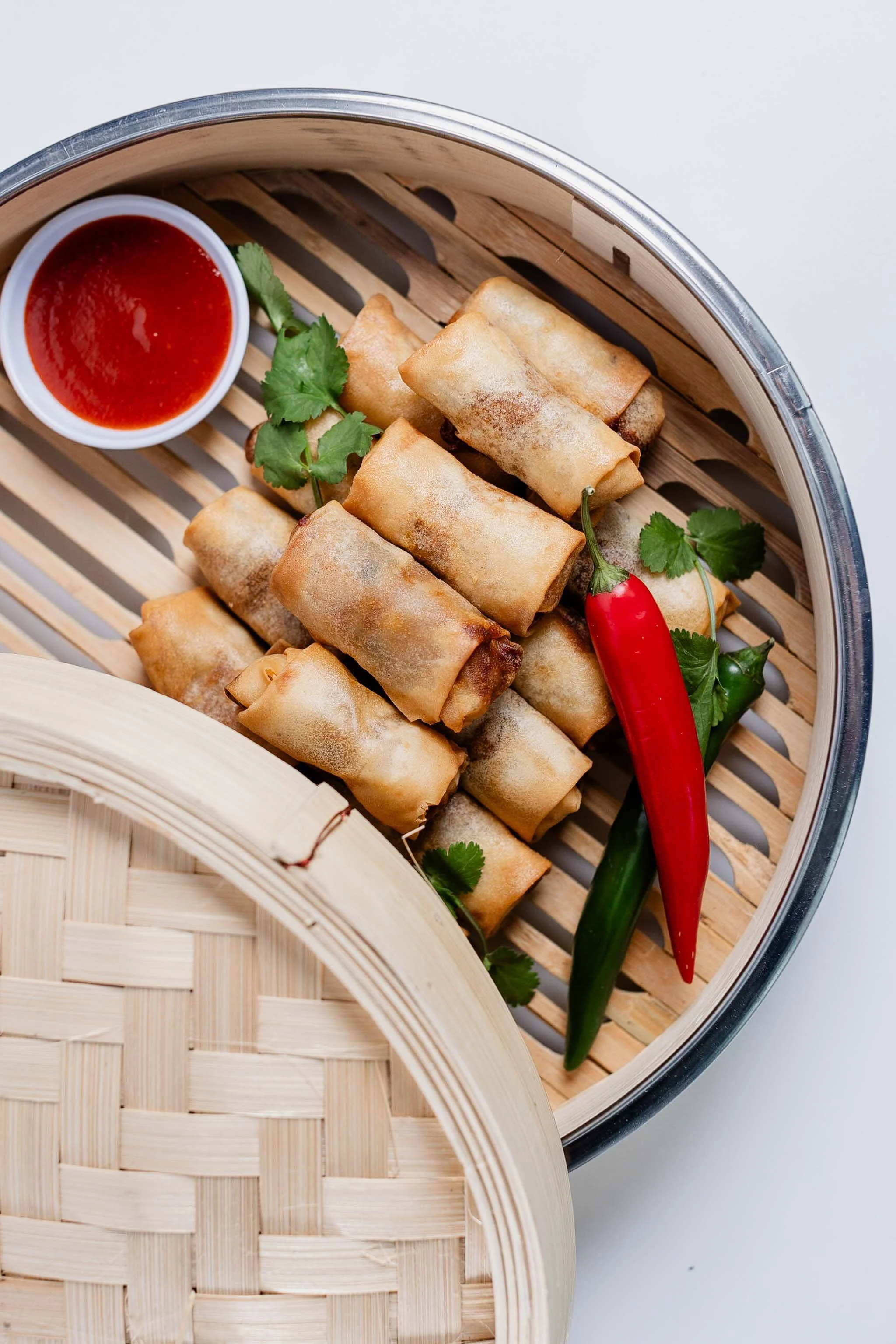 Spring rolls served with a small bowl of red dipping sauce, garnished with cilantro and a red chili pepper in a bamboo steamer basket.