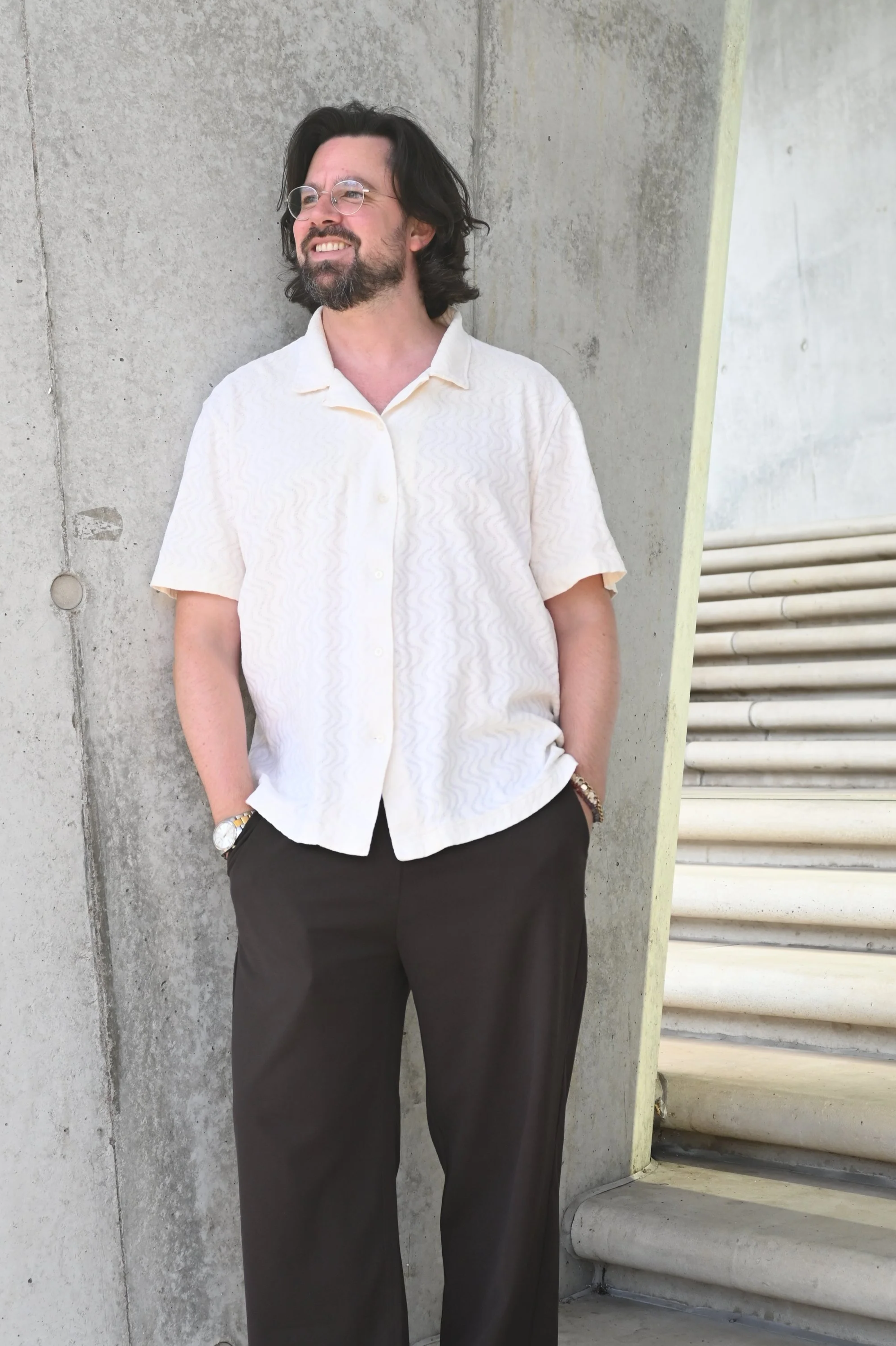 Ein Mann mit dunklen, längeren Haaren und Brille steht lachend vor einer Betonwand auf einer Treppe. Er trägt eine helle, kurzärmelige Hemd und dunkle Hose.
