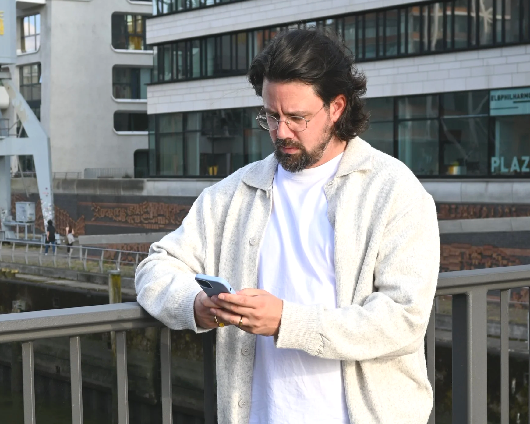 Ein Mann steht auf einer Brücke vor modernen Gebäuden und schaut auf sein Handy.