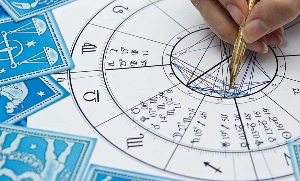 A person writing on an astrological birth chart with a gold pen, surrounded by blue tarot cards.