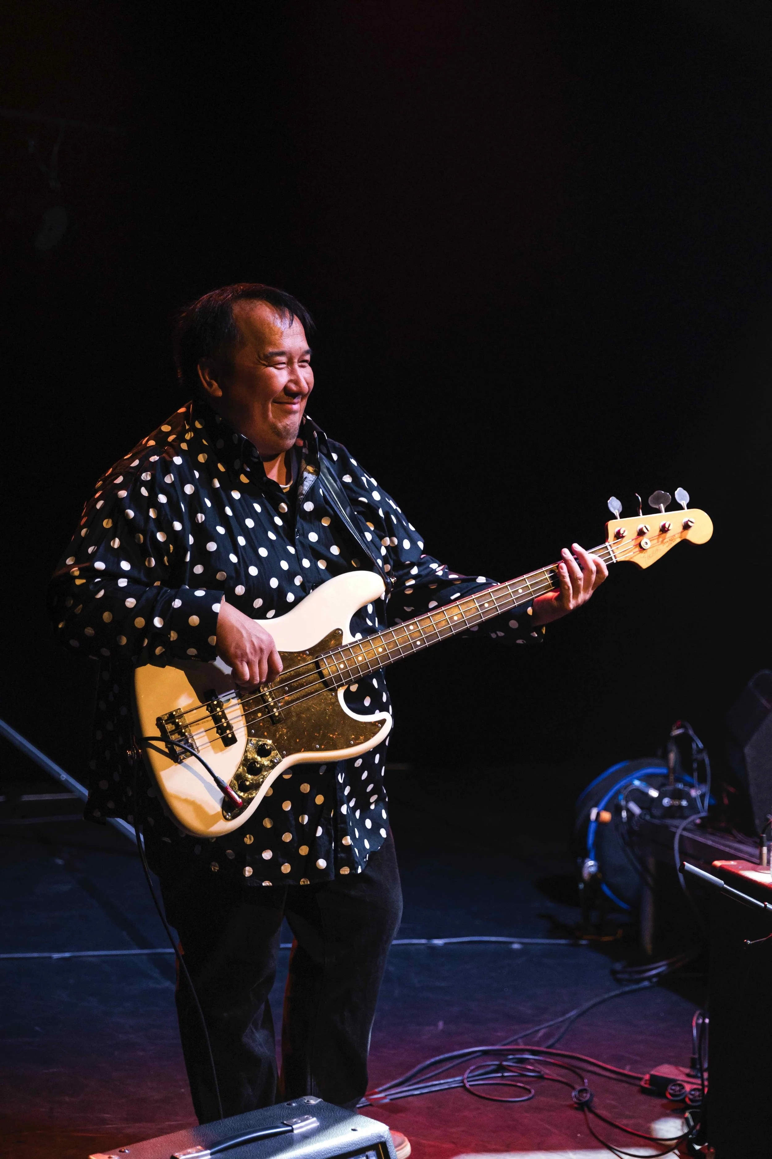 Un homme joue de la basse électrique sur scène, portant une chemise noire à pois blancs, avec un fond sombre.