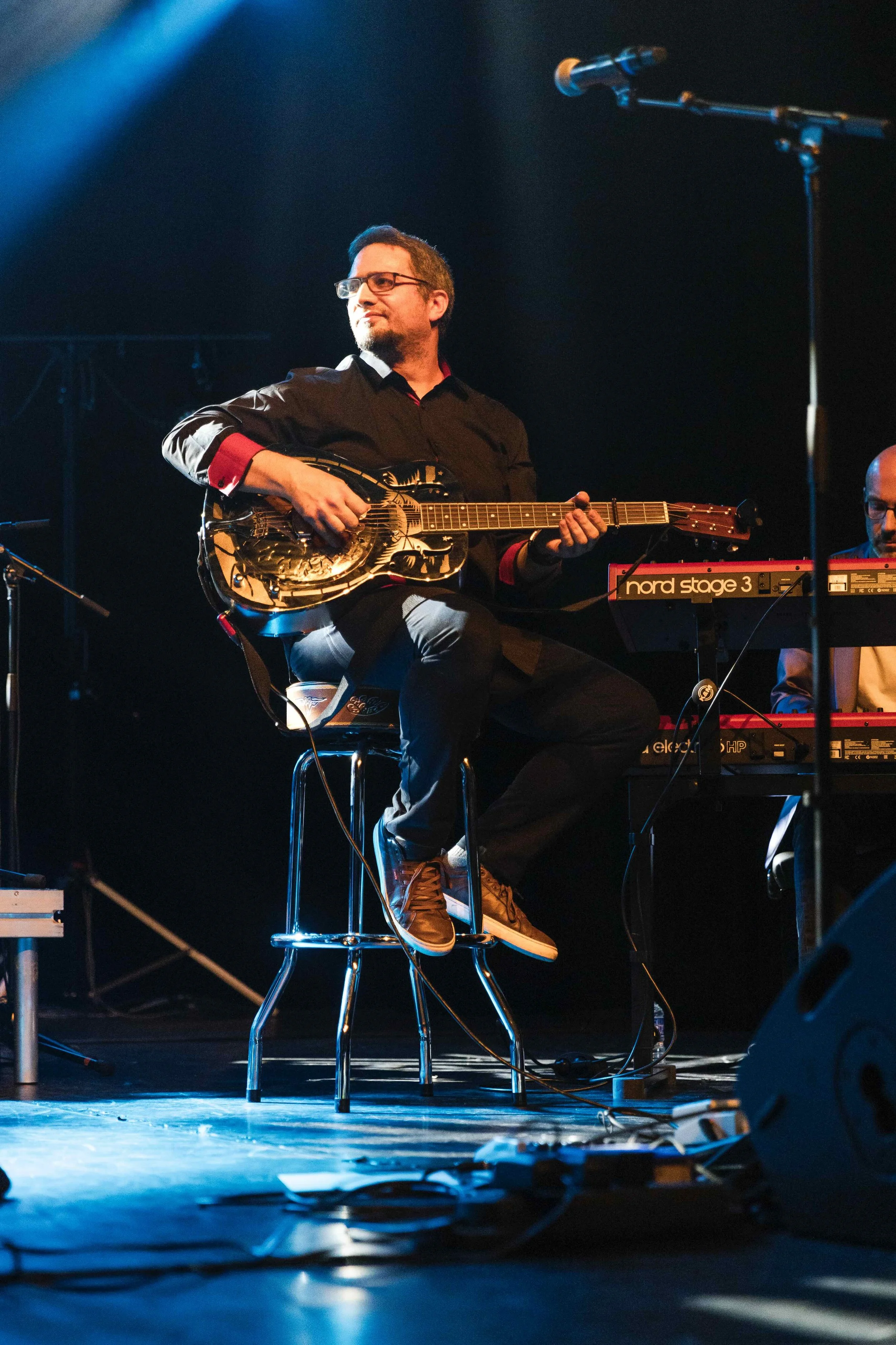 Musicien jouant de la guitare lors d'un concert sur scène, éclairé par des lumières bleues, avec un clavier en arrière-plan.