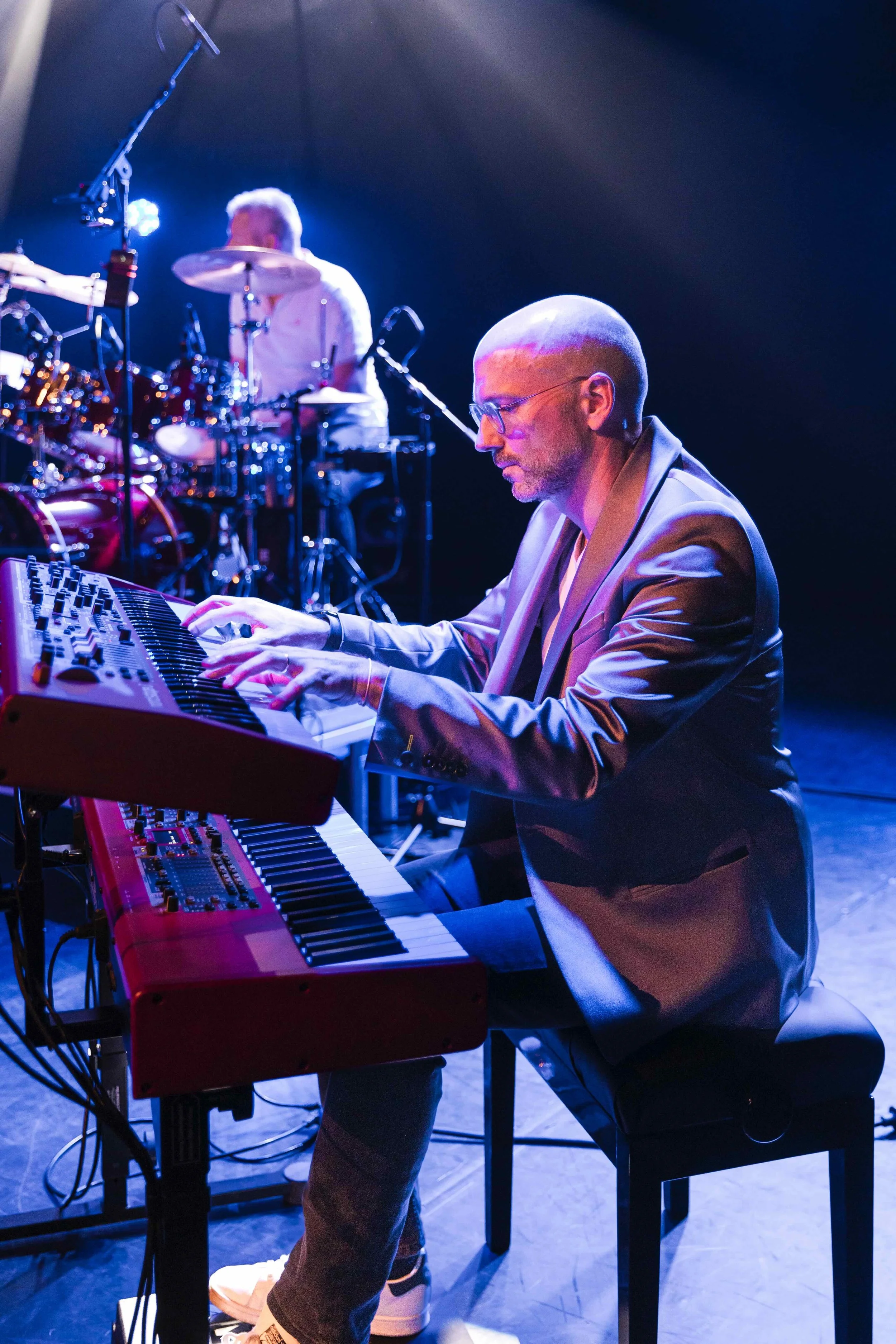 Musiciens jouant du clavier et de la batterie lors d'une performance sur scène, éclairée par des lumières bleues.