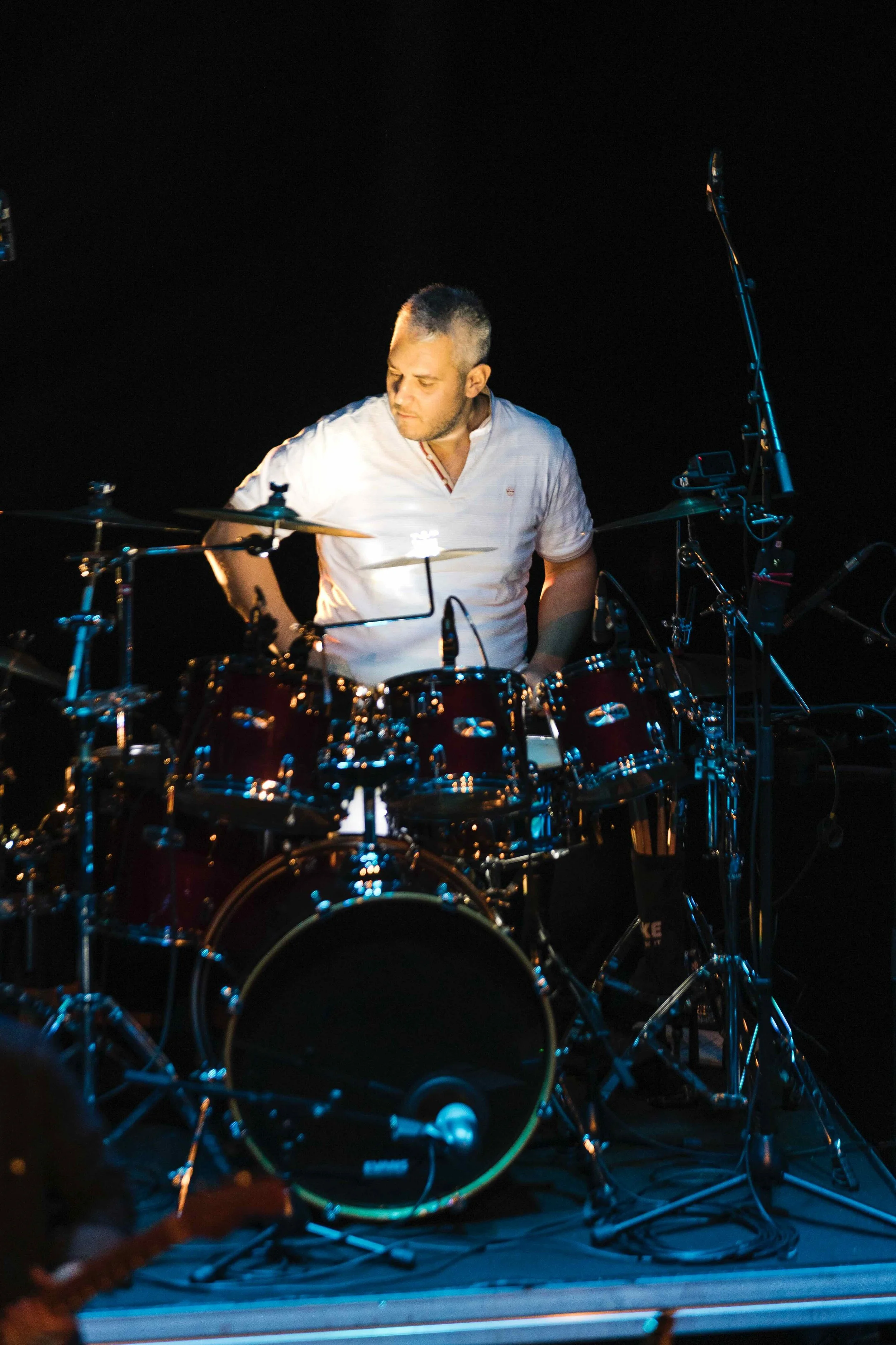Un musicien joue de la batterie sur scène dans un environnement sombre, éclairé par des lumières.