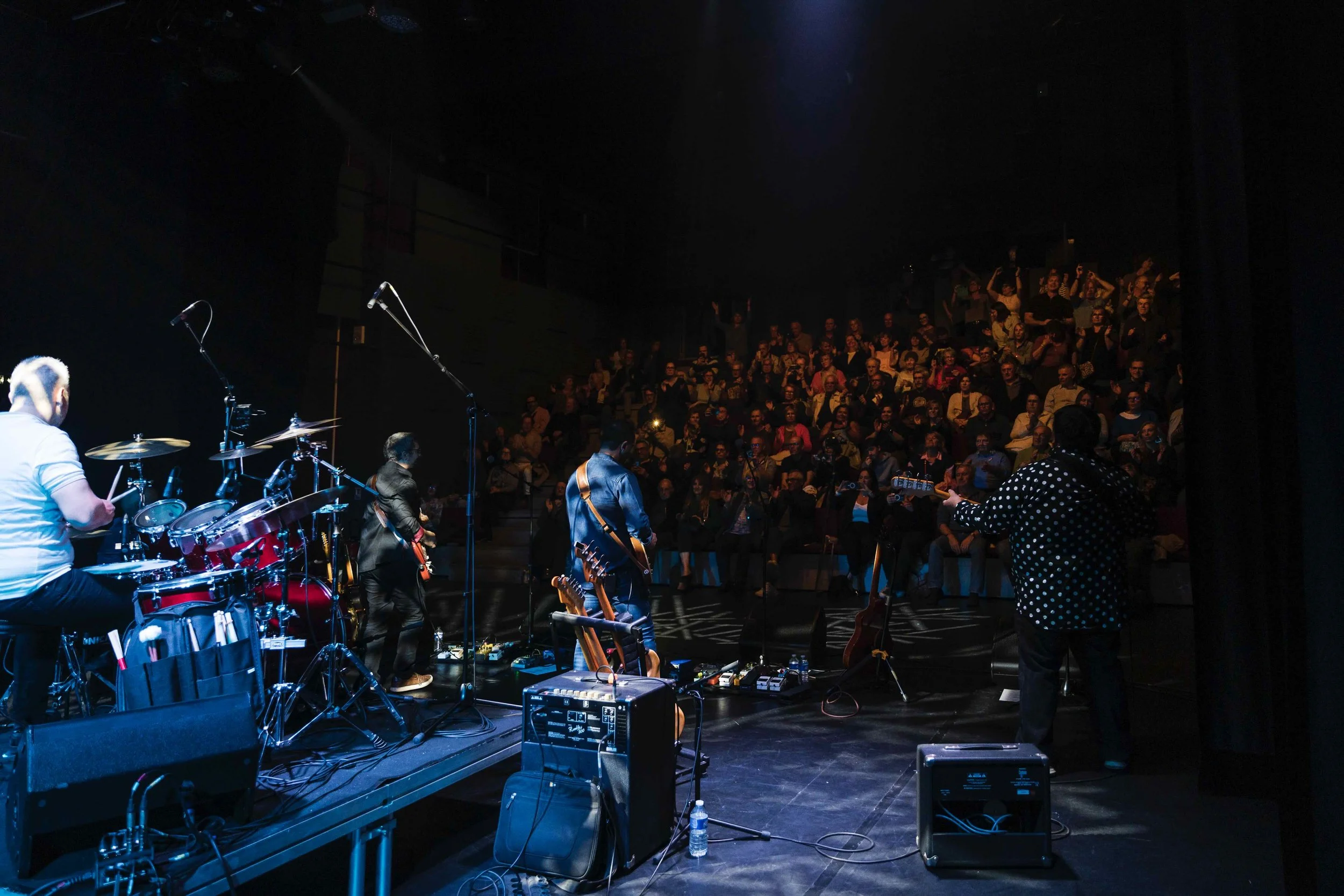 Vue d'une scène de concert avec un groupe de musiciens sur scène et un public assis dans une salle de spectacle sombre.