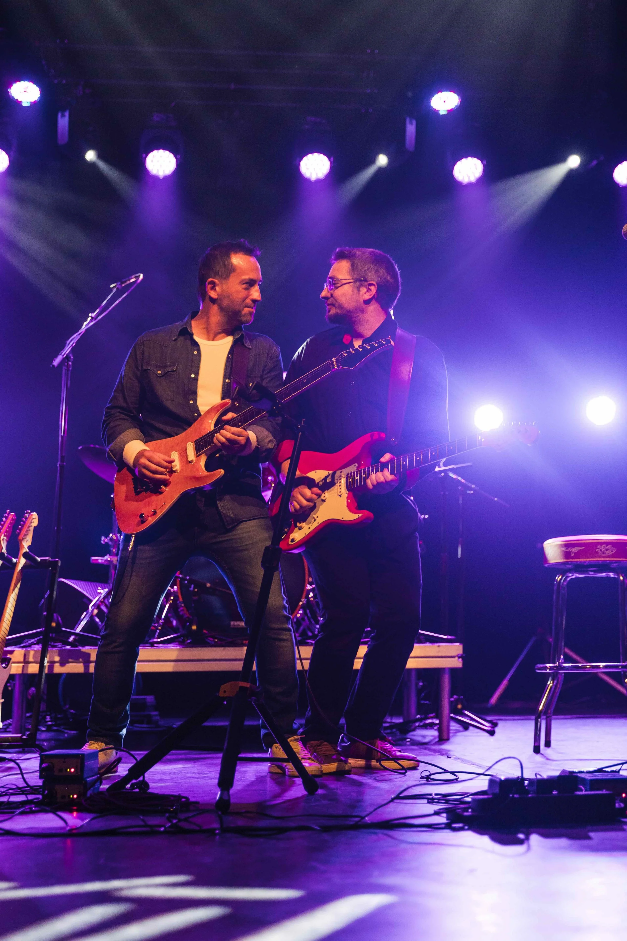 Deux musiciens jouent de la guitare sur une scène éclairée par des lumières violettes, avec du matériel musical autour d'eux.