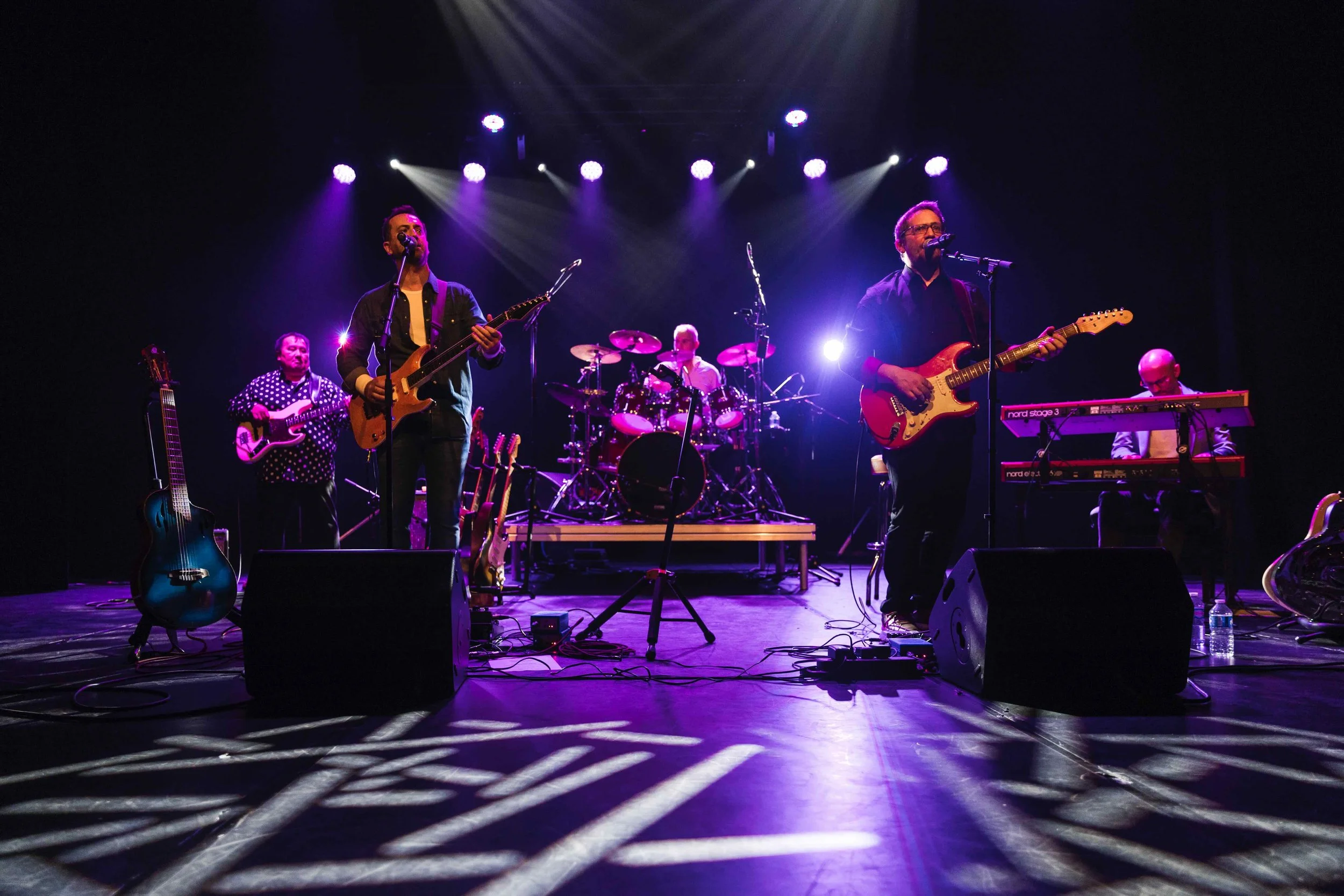 Un groupe de musiciens jouant sur scène avec éclairage violet, comprenant plusieurs guitares, un clavier et une batterie.