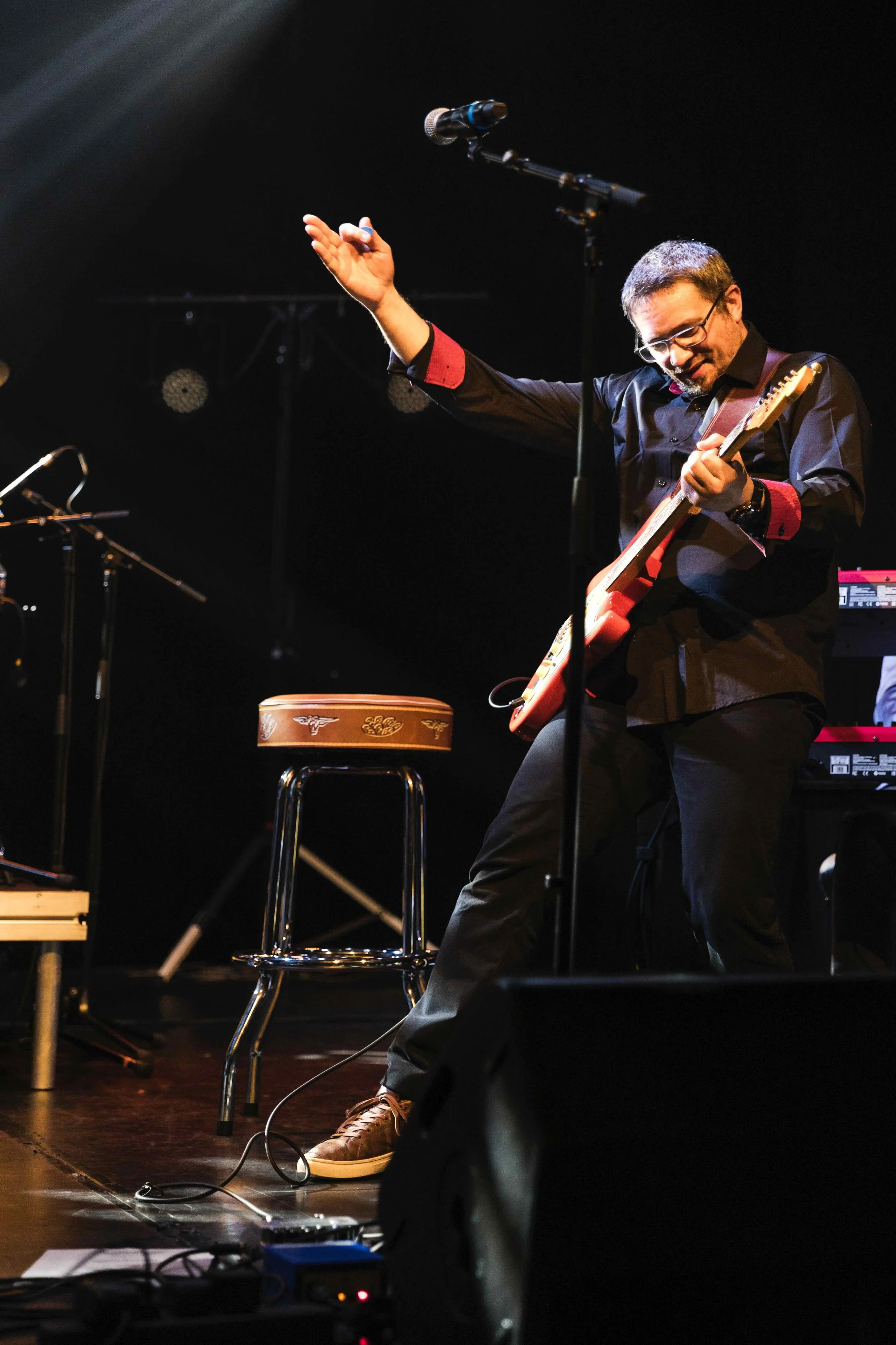 Un homme souriant joue de la guitare électrique sur scène lors d'un concert, avec un micro devant lui et un clavier en arrière-plan.