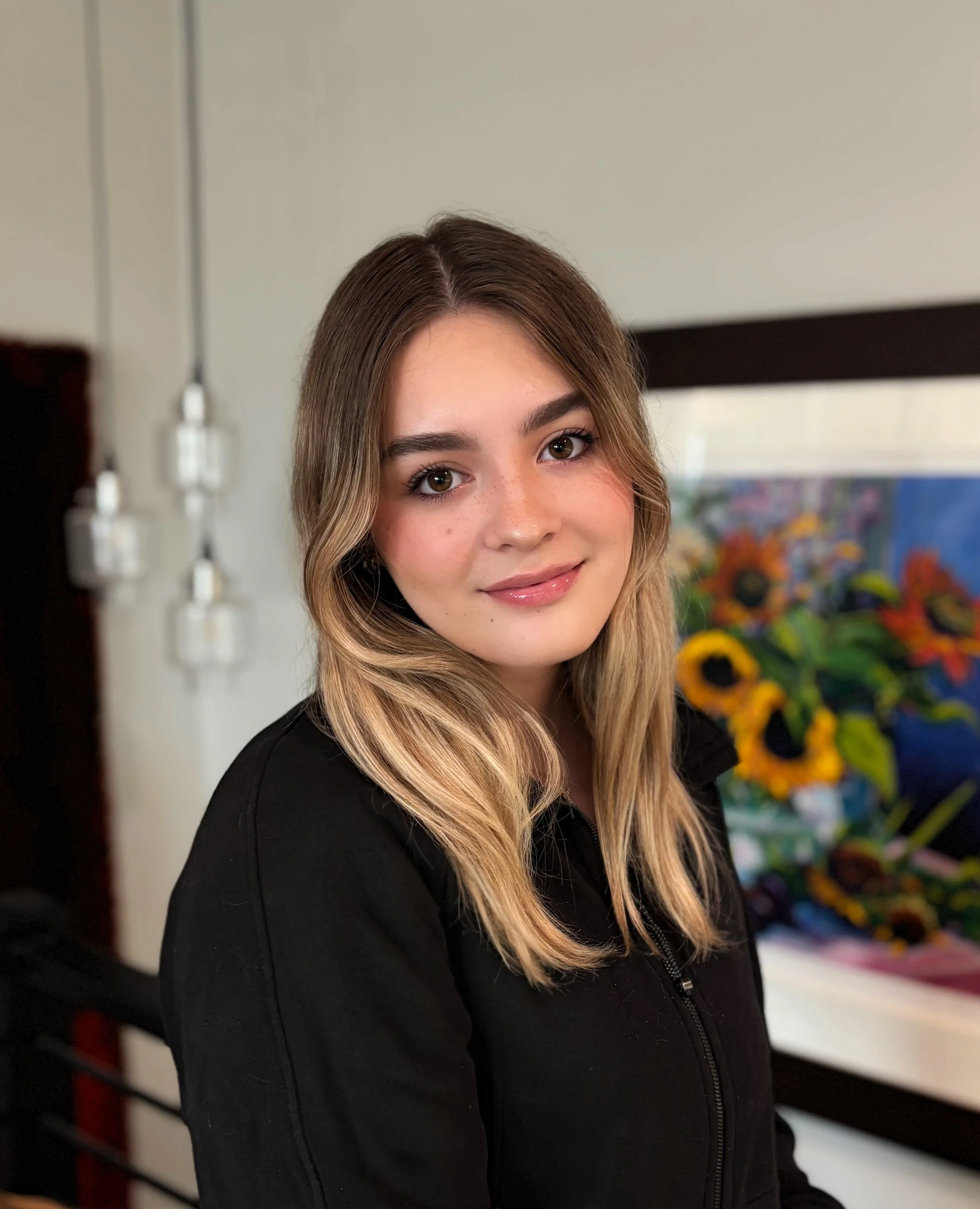 Young woman with shoulder-length blonde hair, smiling, wearing a black jacket, standing in front of a colorful sunflower painting indoors.