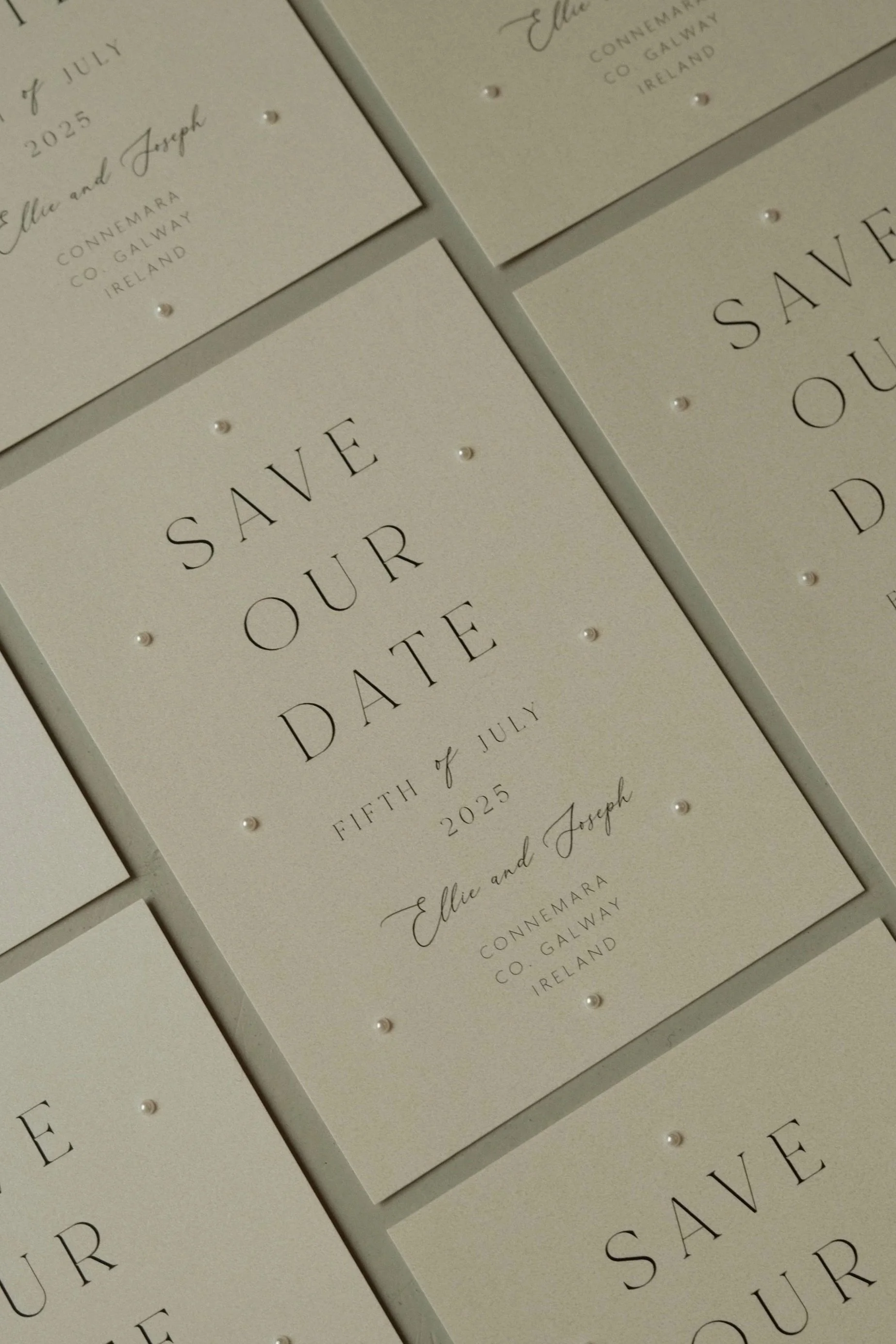 Multiple off-white wedding save-the-date cards with black text and small decorative pearls, arranged in a pattern.