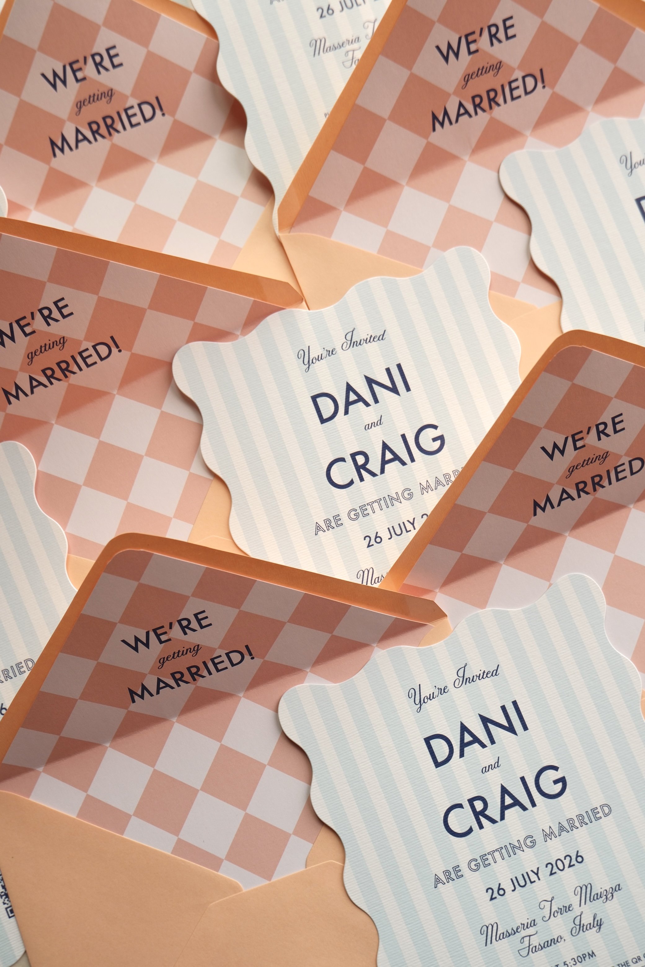 Multiple wedding invitations reading 'You're Invited Dani and Craig are getting married 26 July 2026 Maseria Torre Maizza Fasano Italy' with pink and blue designs, placed in peach envelopes.