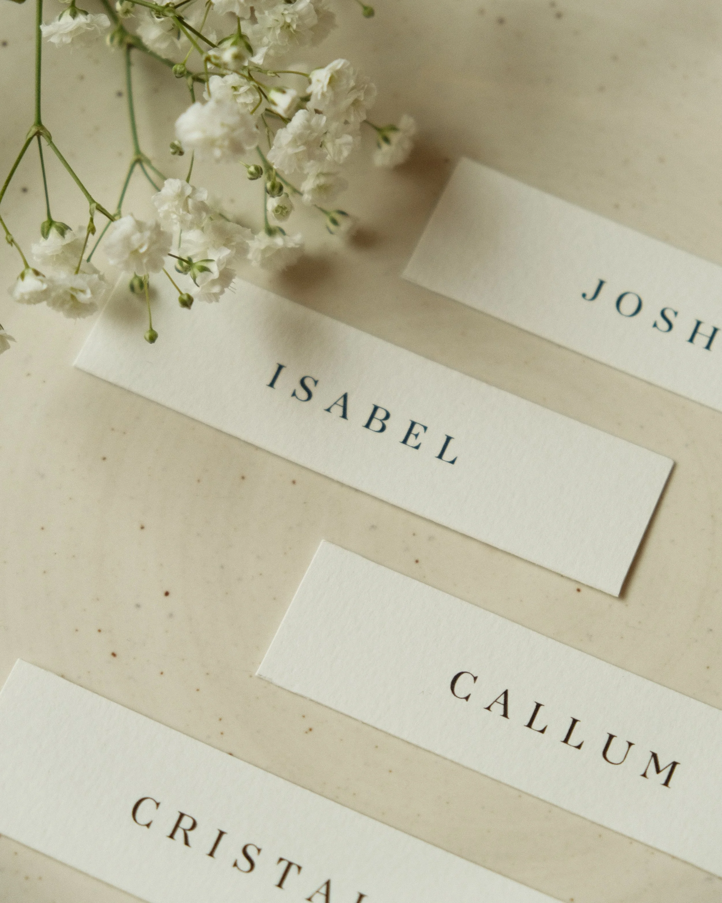 Wedding name cards with the words 'ISABEL', 'JOSH', 'CALLUM', and 'CRISTAL' on a beige surface, with white flowers. For wedding day of stationery items.