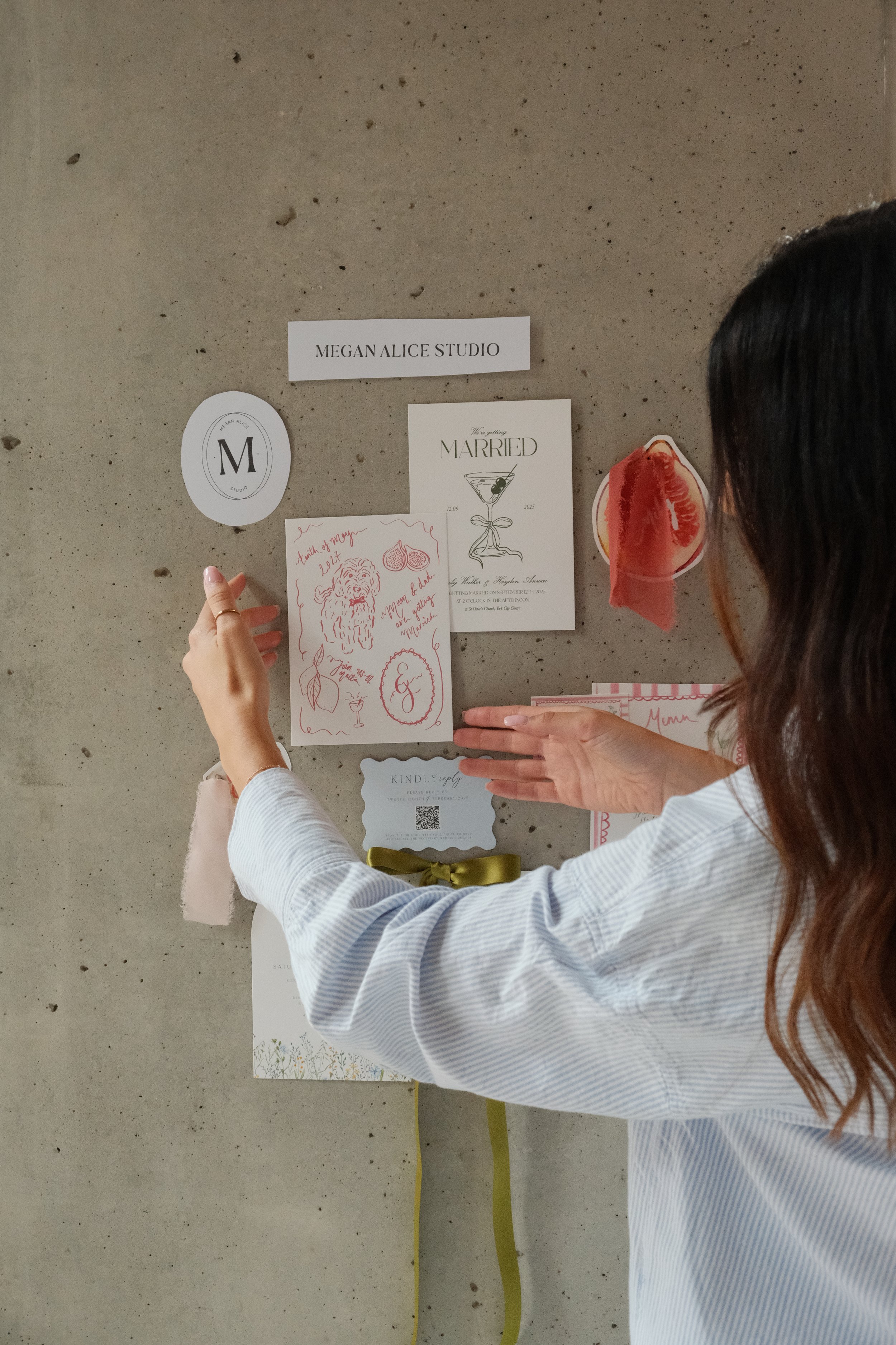 A woman with long brown hair and a white striped shirt arranging a display on a concrete wall with various paper notes, cards, and decorative items, including a wedding announcement and framed initials.
