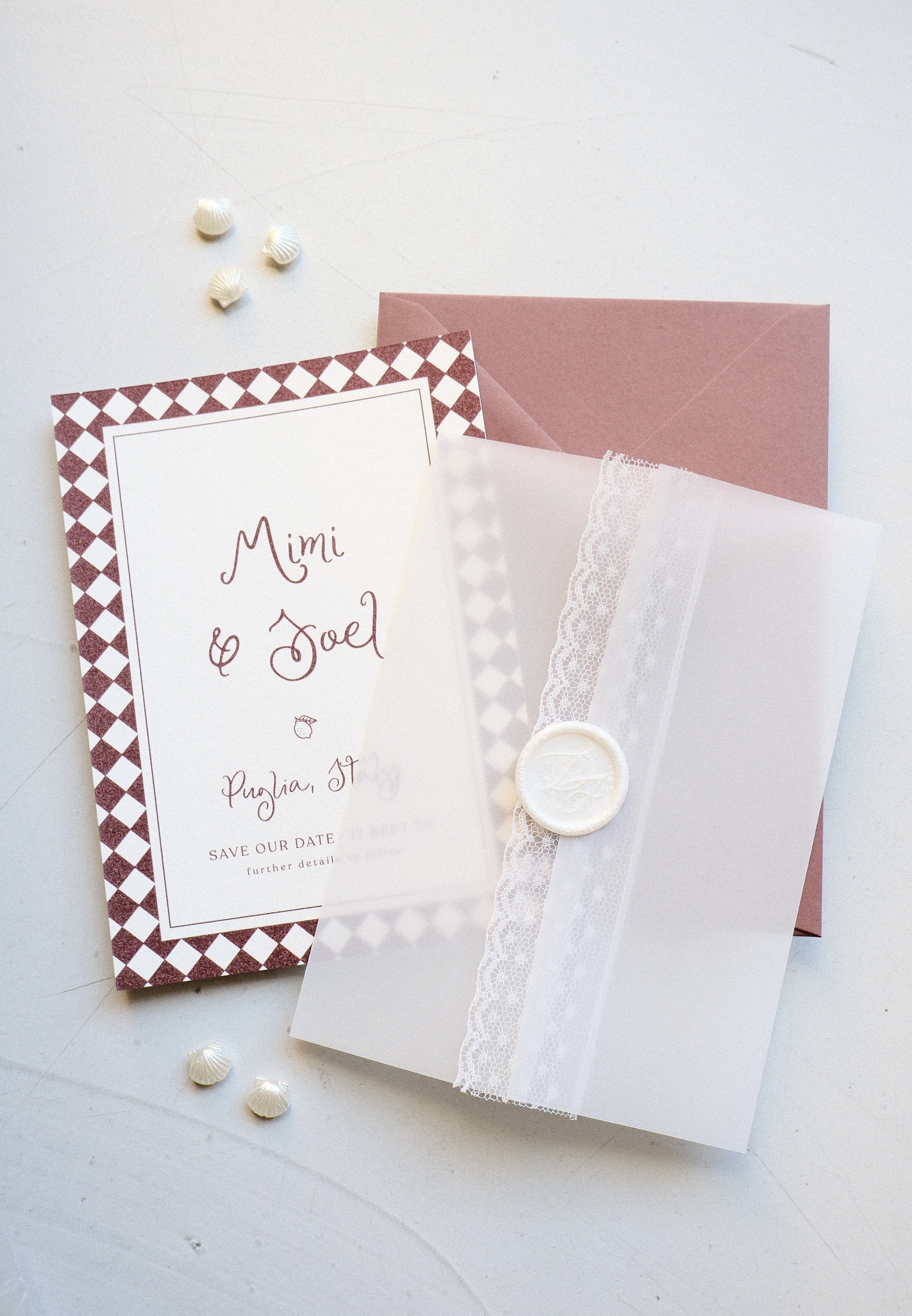 Wedding invitation with lace and wax seal, pink envelope, and seashell decorations.